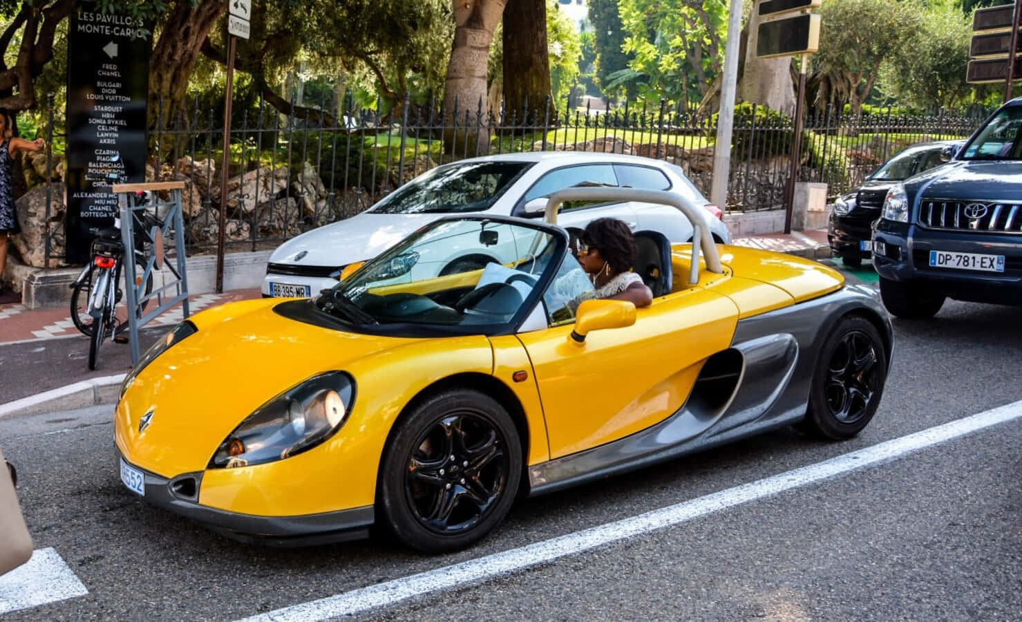 Renault Sport Spider 桌布 壁紙 — 一輛亮黃色、帶黑色裝飾的敞篷跑車在綠樹成蔭的街道上行駛，一名女子駕車經過停放的汽車和一輛自行車。
