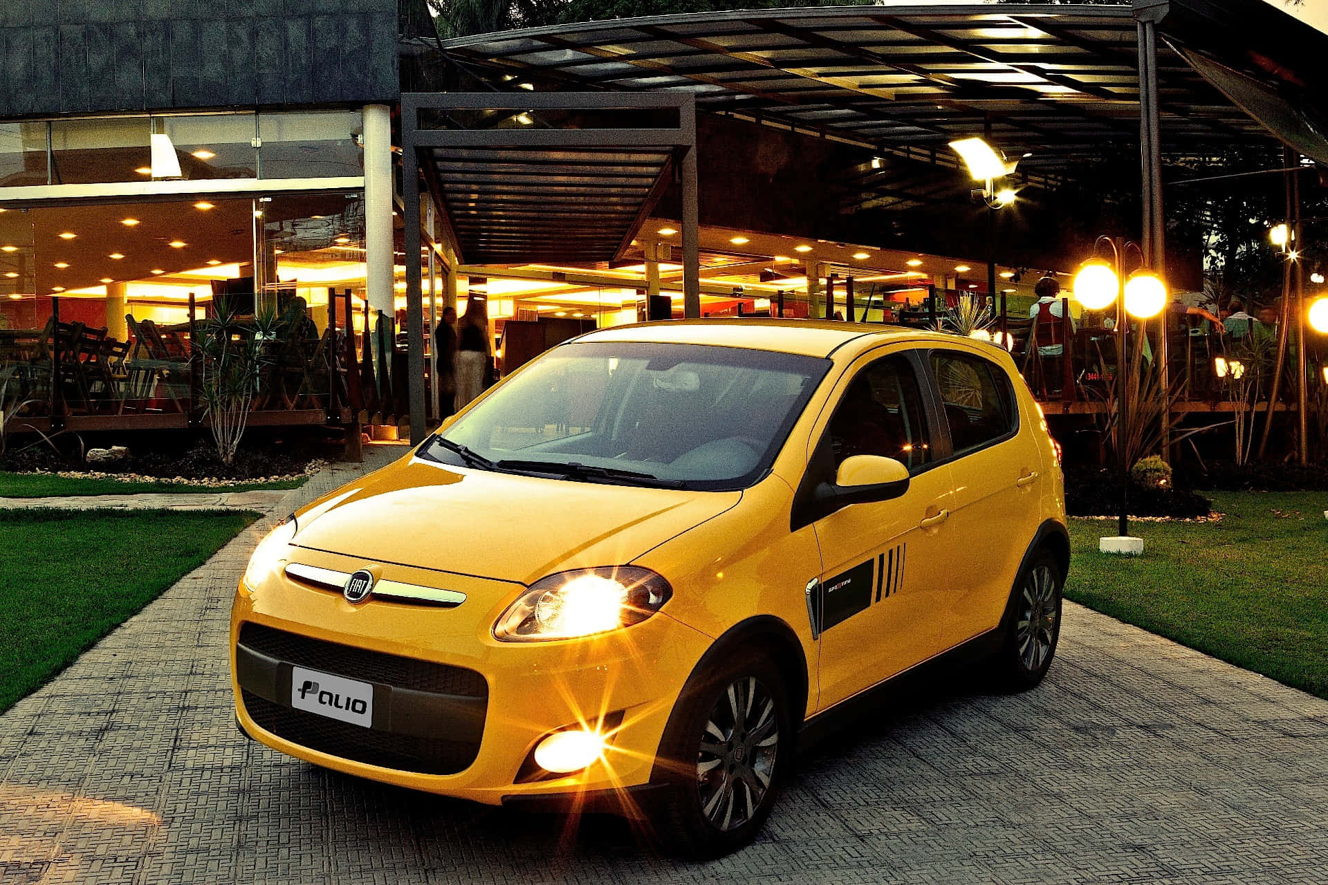 Fiat Palio 桌布 壁紙 — 一輛亮黃色的菲亞特Palio在黃昏時停泊在鋪砌的小路上，背後現代建築散發著溫暖的光芒。