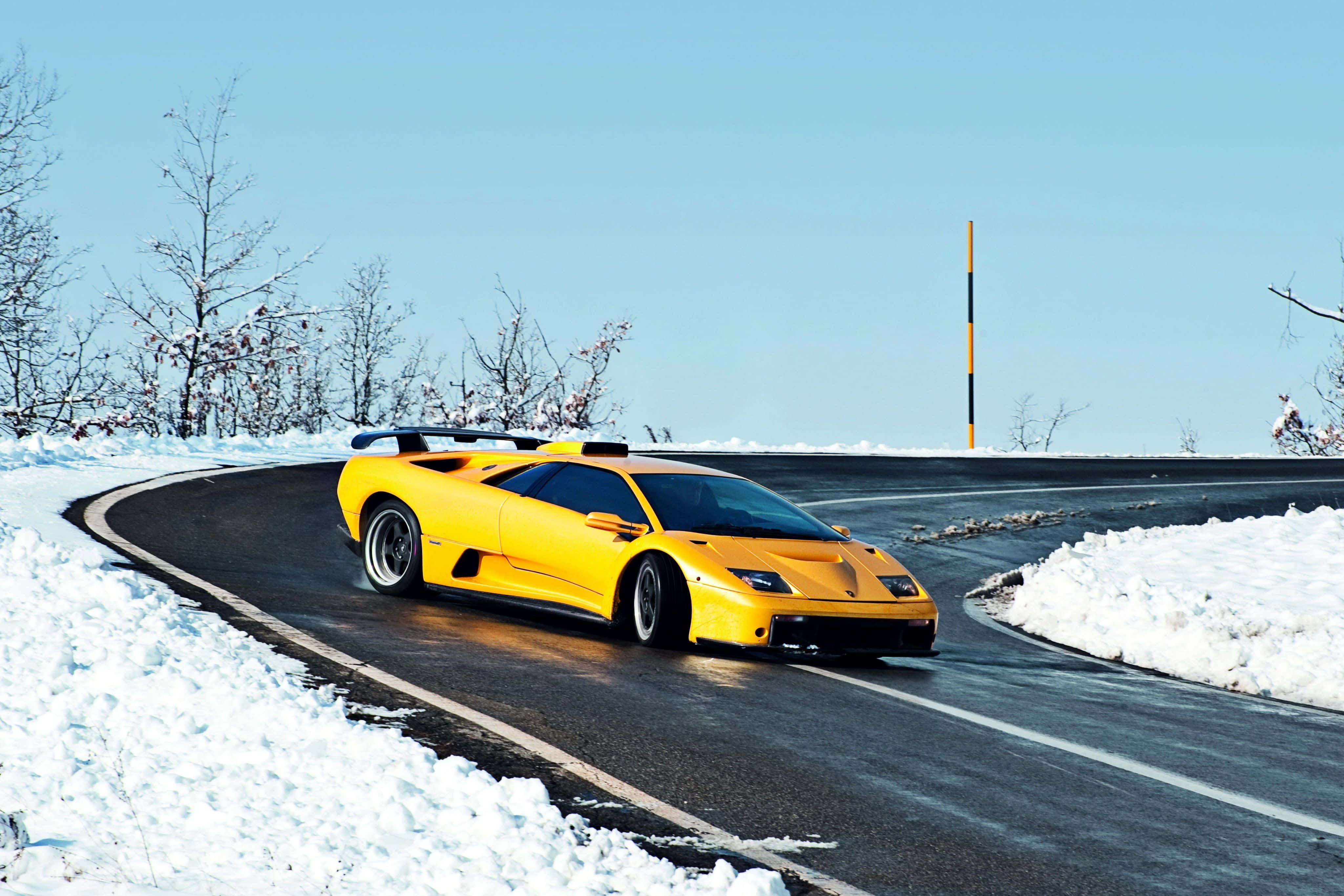 Lamborghini Diablo 桌布 壁紙 — 一輛亮黃色的藍寶堅尼 Murciélago 在晴朗的藍天下，沿著積雪的山路奔馳。