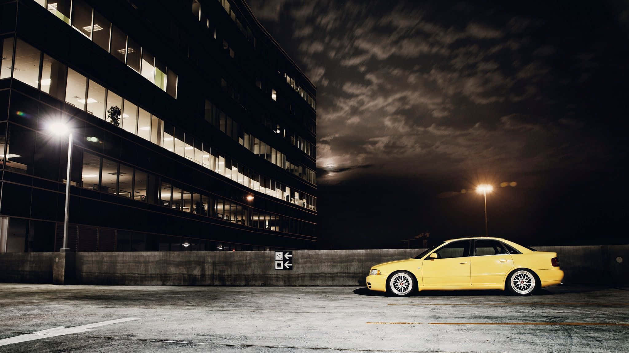 Audi S4 桌布 壁紙 — 一輛亮黃色轎車獨自停泊在夜間昏暗的城市停車場，旁邊是一座帶有發光窗戶的現代建築。