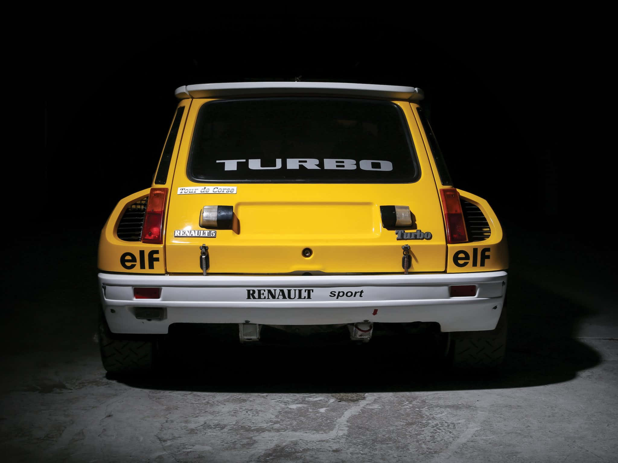 Renault 5 Turbo 桌布 壁紙 — 一輛亮黃色雷諾5 Turbo跑車的尾部在黑暗背景下被照亮，展示其標誌性賽車貼紙和侵略性設計。