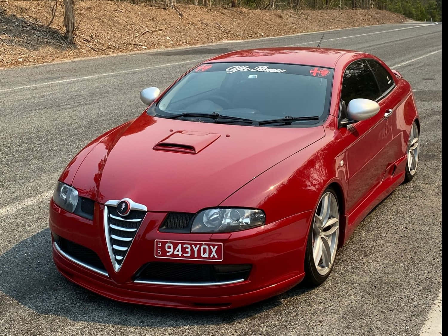 Alfa Romeo Gt 桌布 壁紙 — 一輛亮麗的紅色愛快羅密歐跑車停泊在陽光照耀的瀝青路上，背景有乾草和樹木。