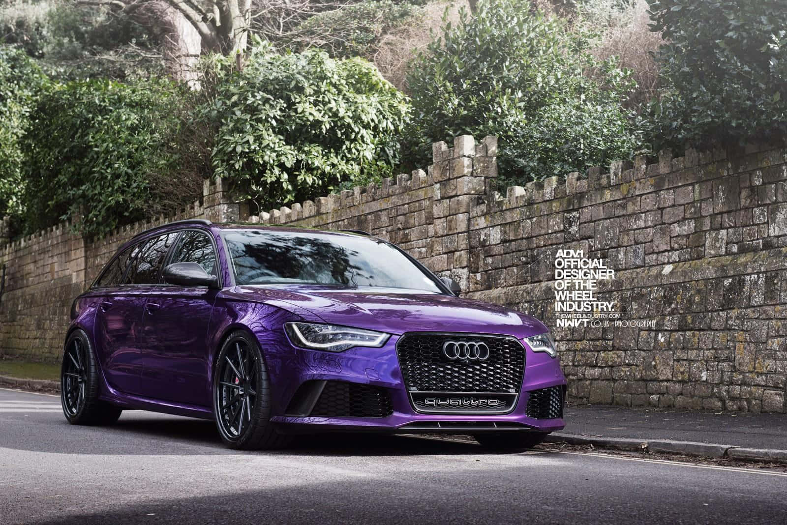 Audi Rs6 桌布 壁紙 — 一輛亮麗的紫色 Audi RS6 Avant 運動旅行車停泊在街上，旁邊是石牆和綠色樹葉。