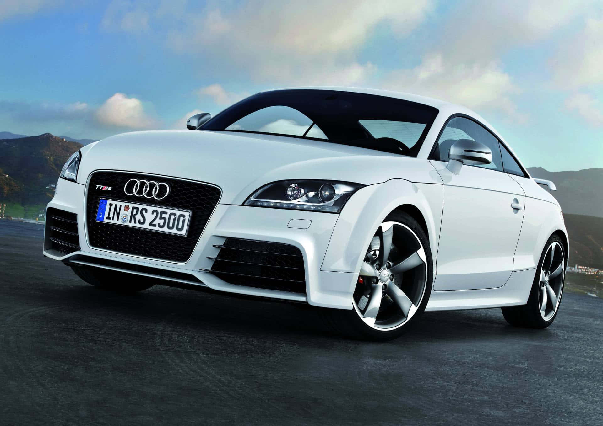 Audi Tt Rs 桌布 壁紙 — 一輛流線型白色奧迪TT RS轎跑車在明亮的日光下，以起伏的丘陵和部分多雲的天空為背景閃閃發光。