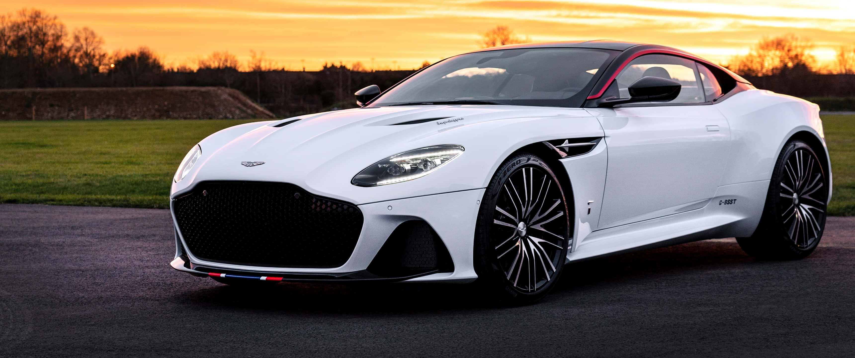 Aston Martin Dbs Superleggera 桌布 壁紙 — 一輛流線型白色Aston Martin DBS Superleggera在日落時分停在柏油路上，背後是金色天空和綠色田野。