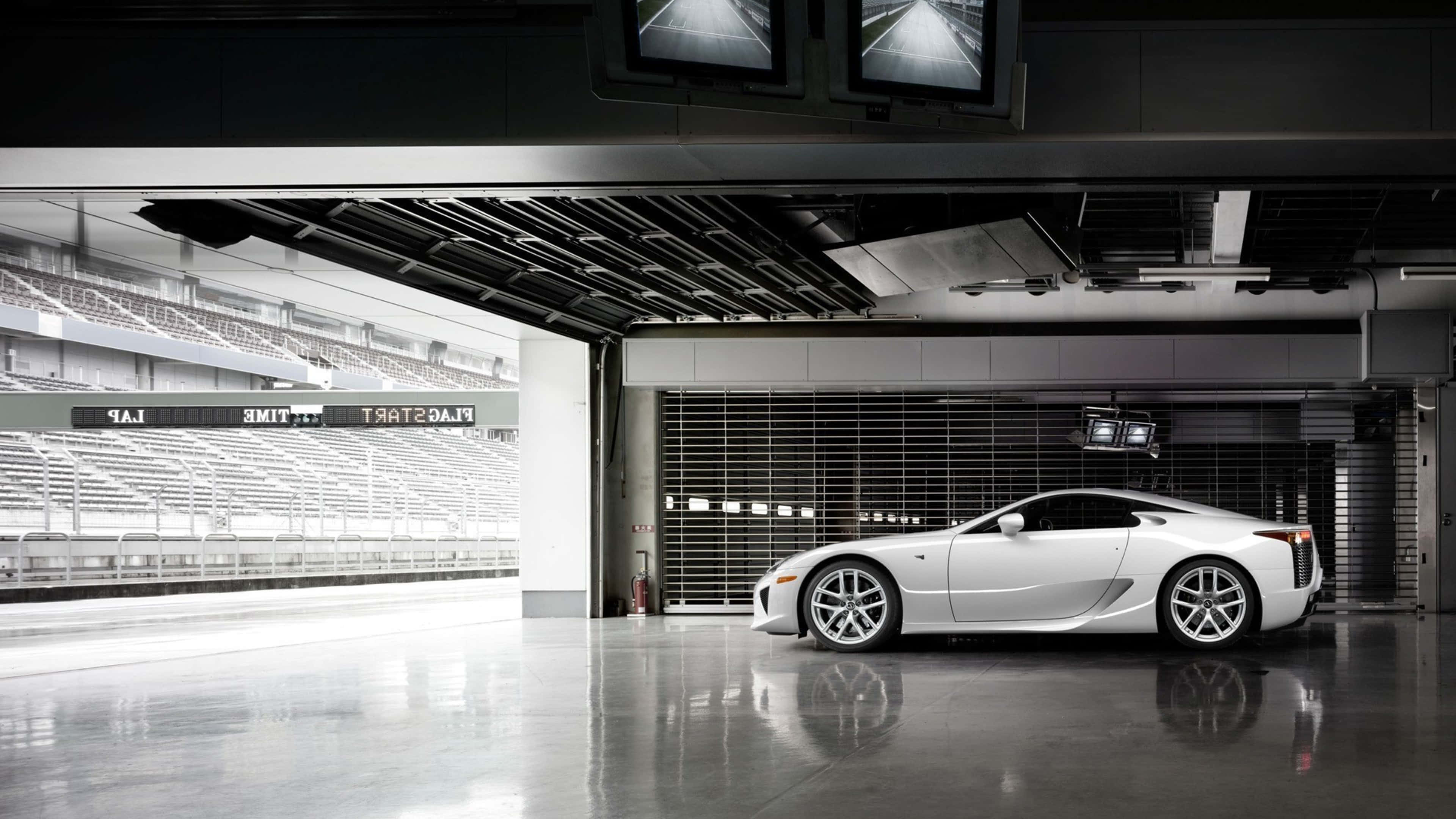Lexus Lfa 桌布 壁紙 — 一輛流線型白色跑車在現代車庫的工業燈光下閃閃發光，旁邊是空無一人的賽車場看台。