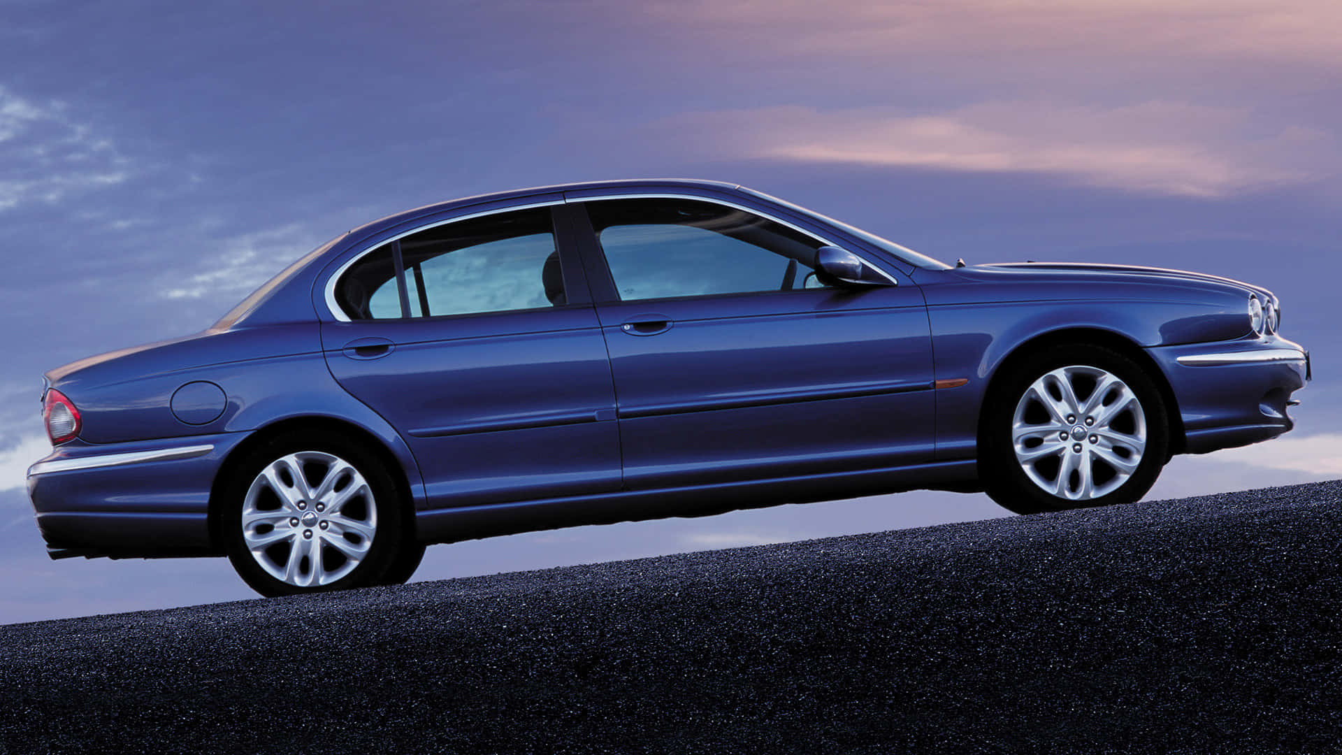 Jaguar X-type 桌布 壁紙 — 一輛流線型的藍色積架轎車在暮色天空下，沿著黑暗的道路滑行，天空中有柔和的紫色和粉紅色雲彩。
