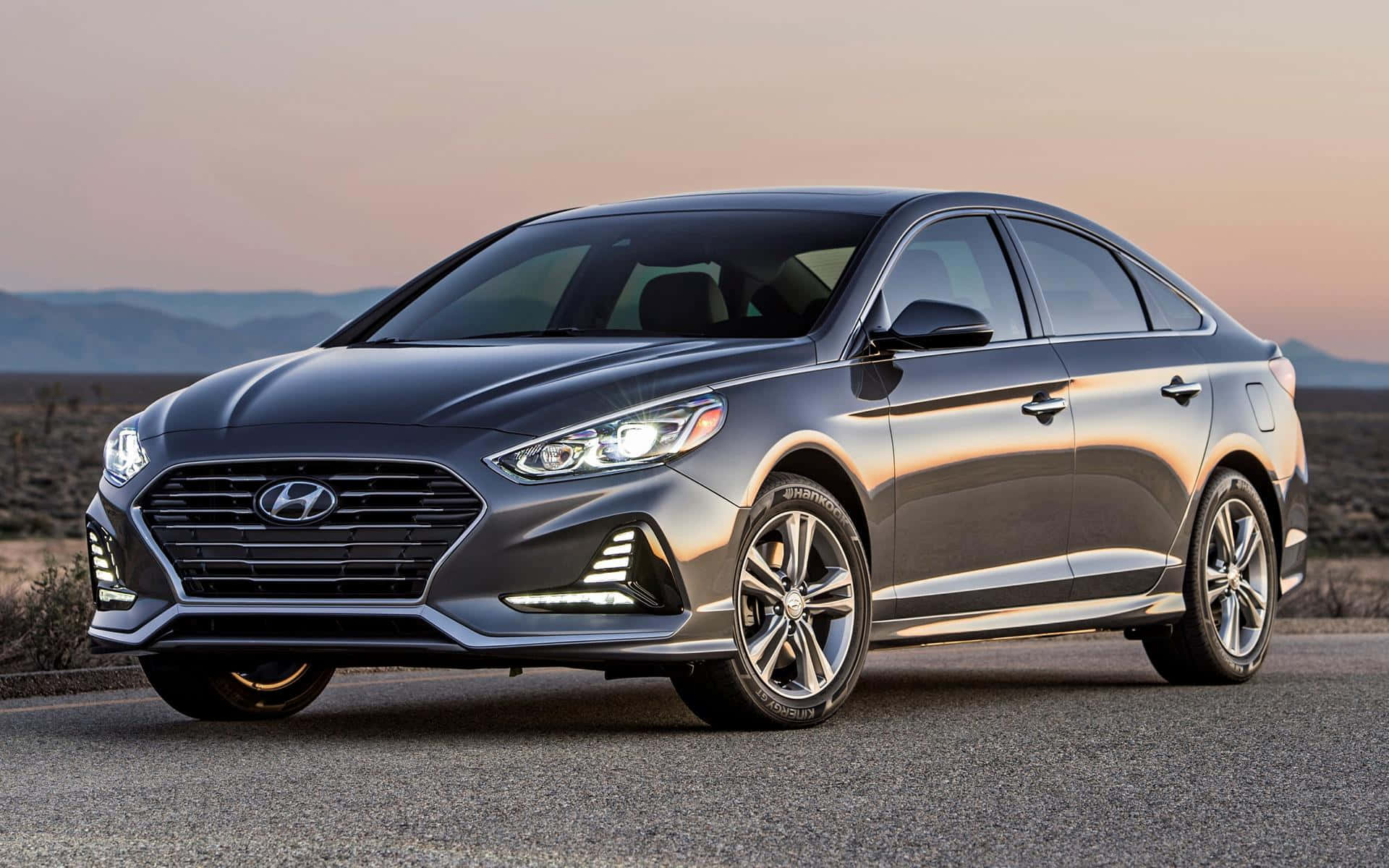 Hyundai Sonata 桌布 壁紙 — 一輛流線型灰色現代Sonata轎車在沙漠公路上，夕陽餘暉下閃耀，遠山為背景。