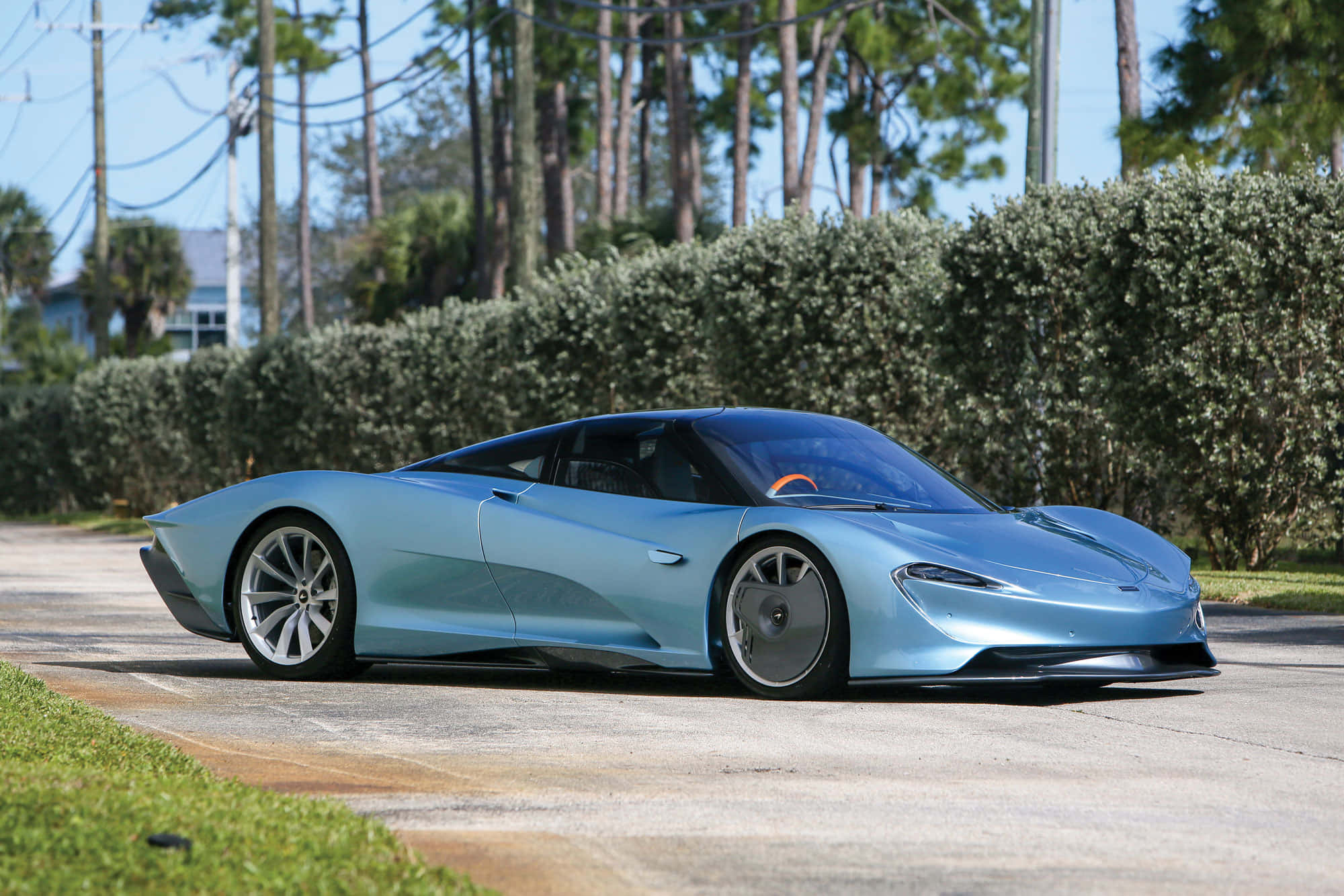 Mclaren Speedtail 桌布 壁紙 — 一輛流線型淺藍色McLaren Speedtail跑車停放在鋪砌道路上，旁邊有綠色籬笆和高大的樹木，天空清澈湛藍。