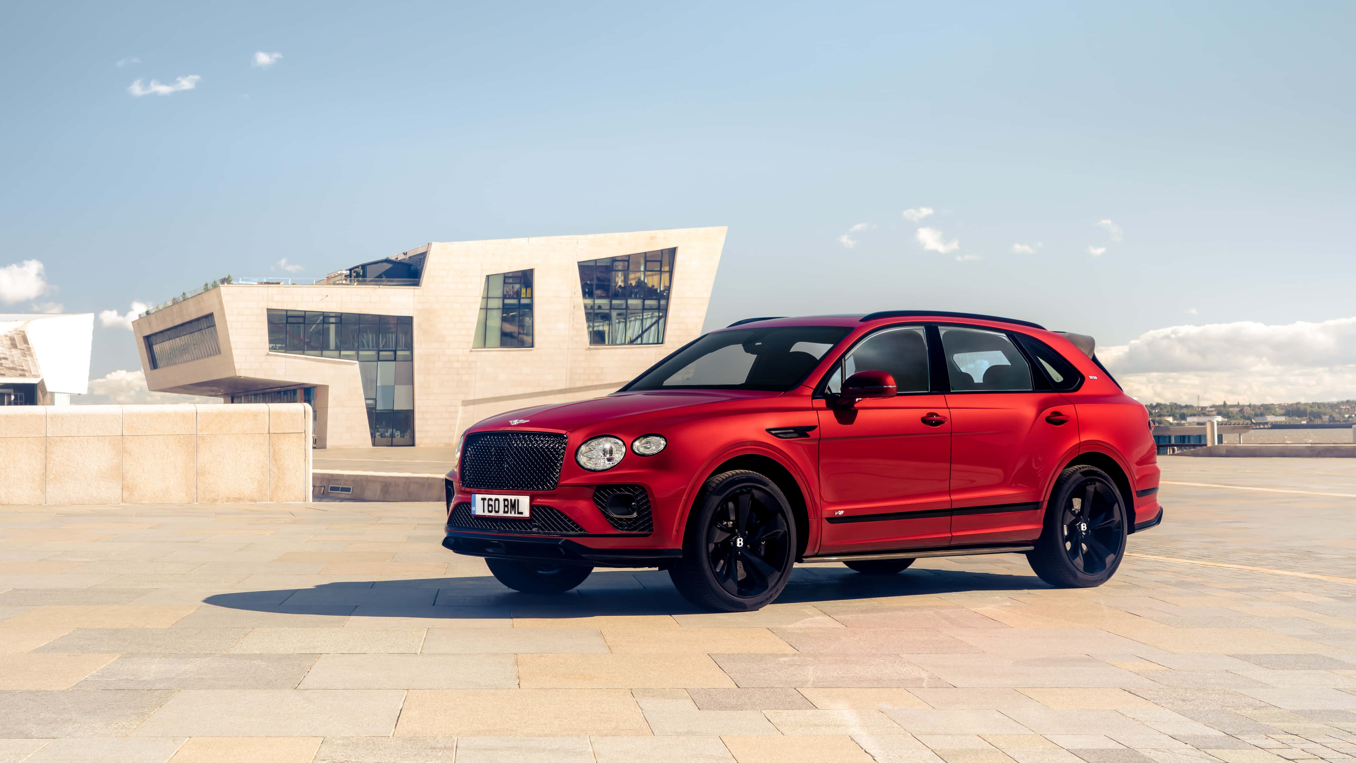 Bentley Bentayga 桌布 壁紙 — 一輛搶眼的紅色賓利Bentayga SUV停放在石頭廣場上，旁邊是現代米色建築，天空清澈湛藍。