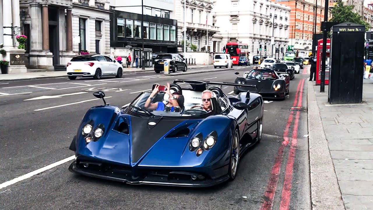 Pagani Zonda Hp Barchetta 桌布 壁紙 — 一輛搶眼的藍黑 Pagani Huayra 跑車在繁忙的倫敦街道上行駛，車上兩名微笑的乘客正在揮手。
