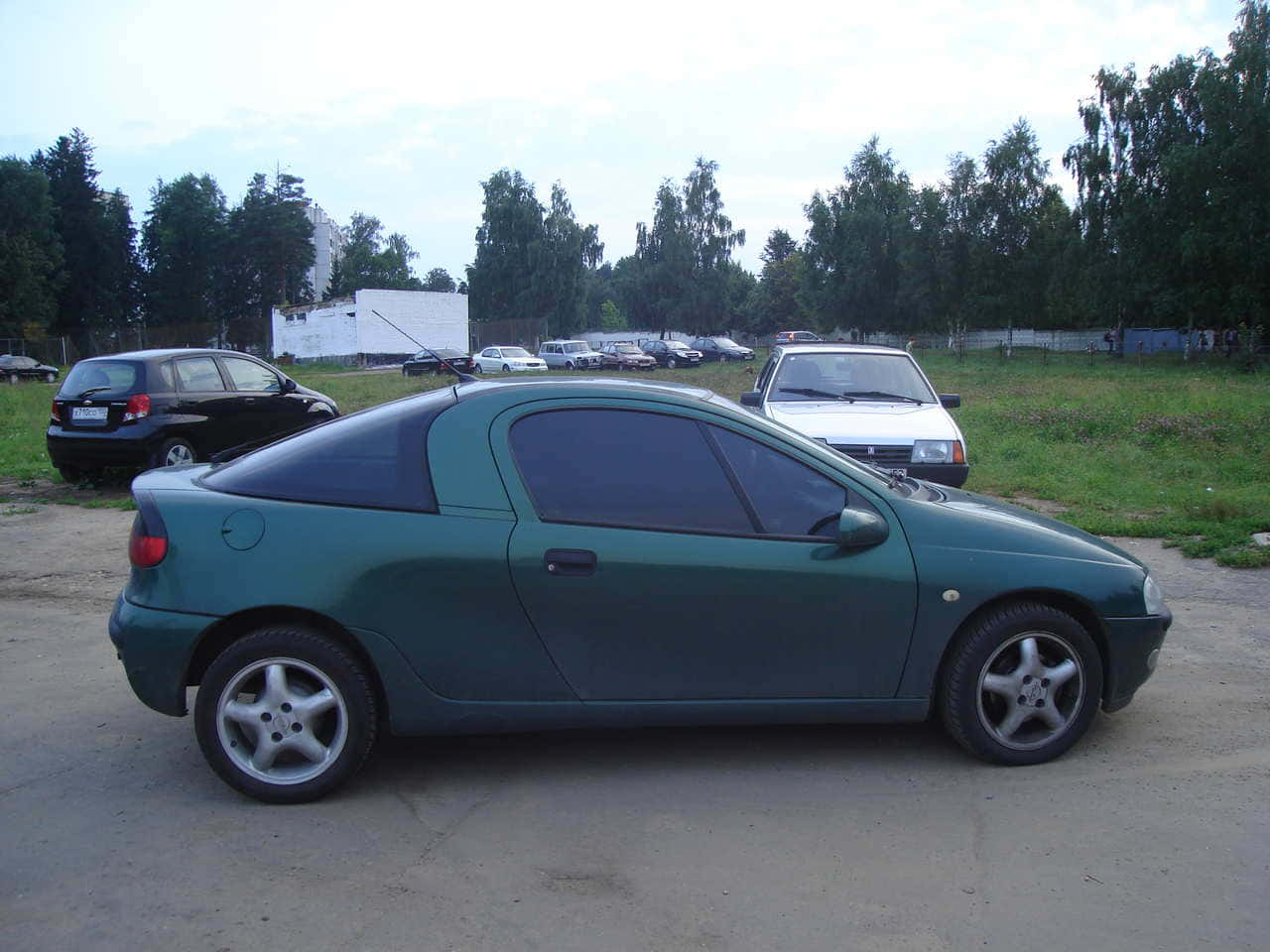 Opel Tigra 桌布 壁紙 — 一輛青綠色雙門轎跑車停放在碎石地上，背景有樹木和其他車輛，天空多雲。