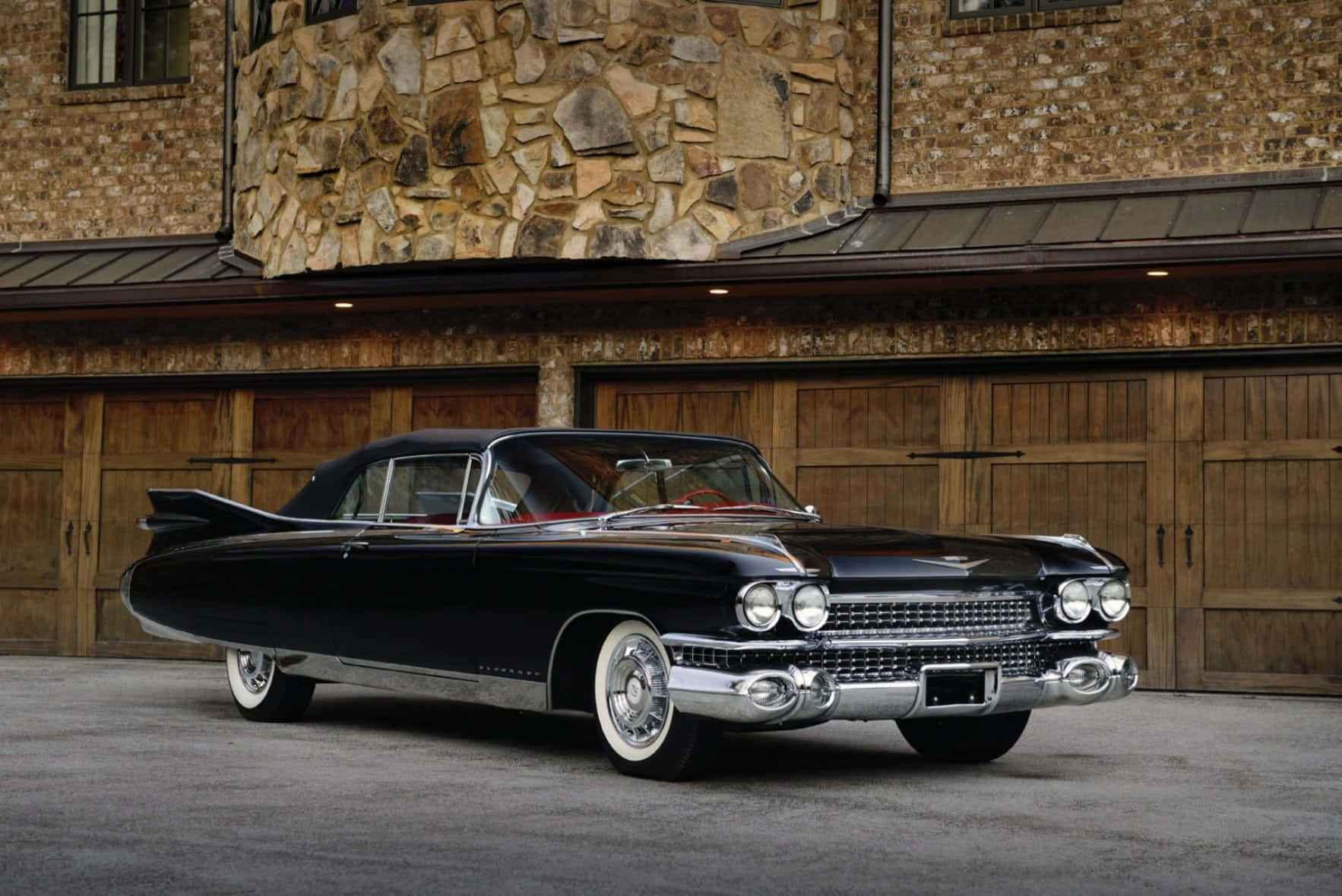 Cadillac Eldorado 桌布 壁紙 — 一輛閃亮的黑色1959年凱迪拉克，帶有鍍鉻裝飾和白色輪胎，停在質樸的石木車庫前。