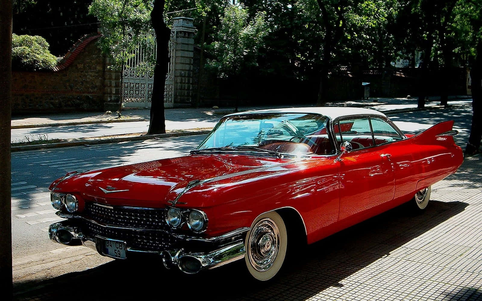 Cadillac Eldorado 桌布 壁紙 — 一輛閃亮的紅色1959年凱迪拉克Eldorado Biarritz在明亮的陽光下停泊在綠樹成蔭的鵝卵石街道上閃閃發光。