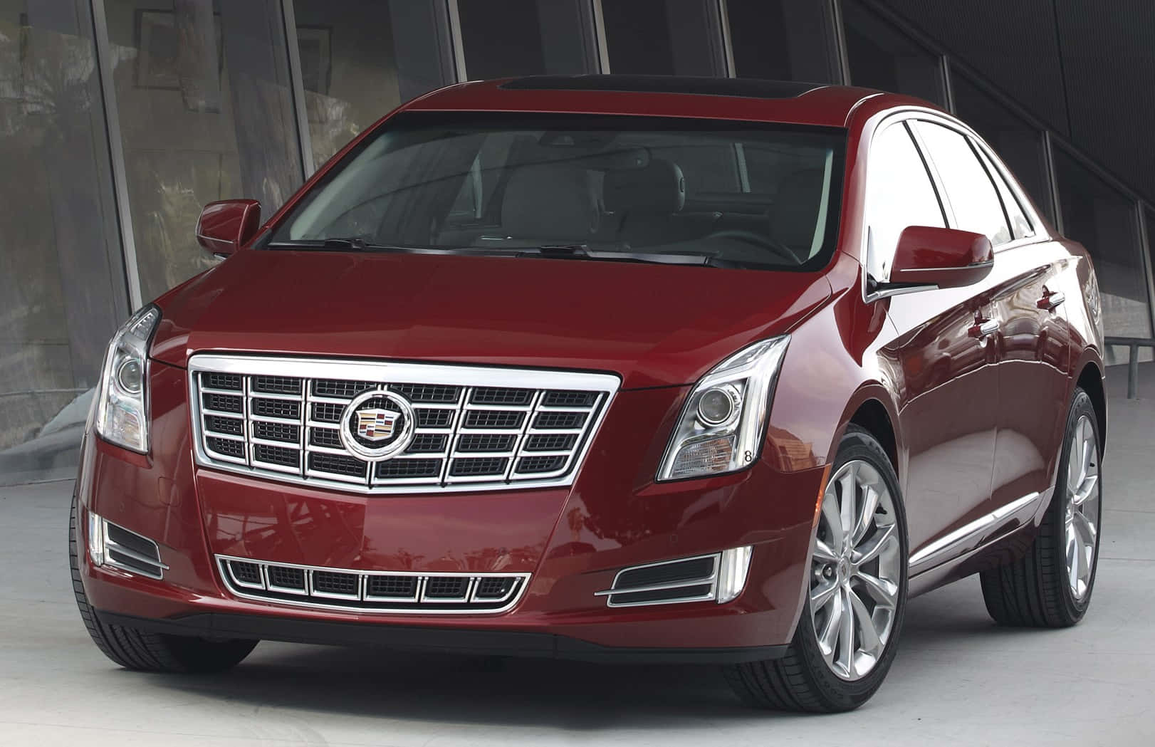 Cadillac Xts 桌布 壁紙 — 一輛閃亮的紅色凱迪拉克轎車在明亮的攝影棚燈光下，以現代玻璃建築為背景閃閃發光。
