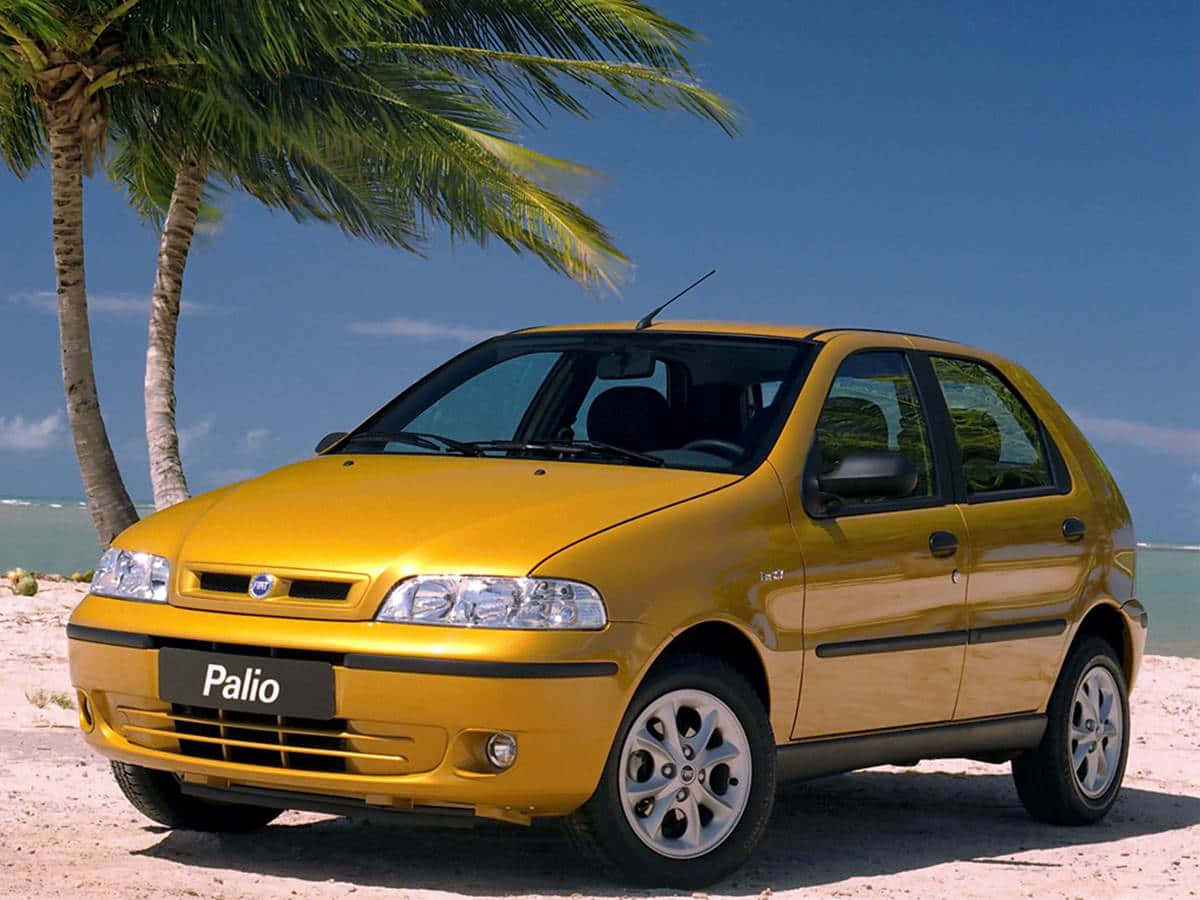 Fiat Palio 桌布 壁紙 — 一輛閃亮的黃色Fiat Palio停泊在棕櫚樹下的沙灘上，背景是海洋和藍天。