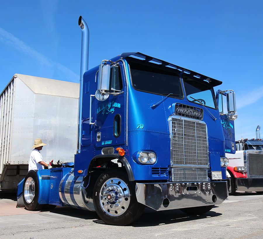 Kenworth T604 桌布 壁紙 — 一輛閃亮的藍色 Freightliner 半掛卡車停在晴朗的藍天下，附近有一名戴帽子的男子正在工作。