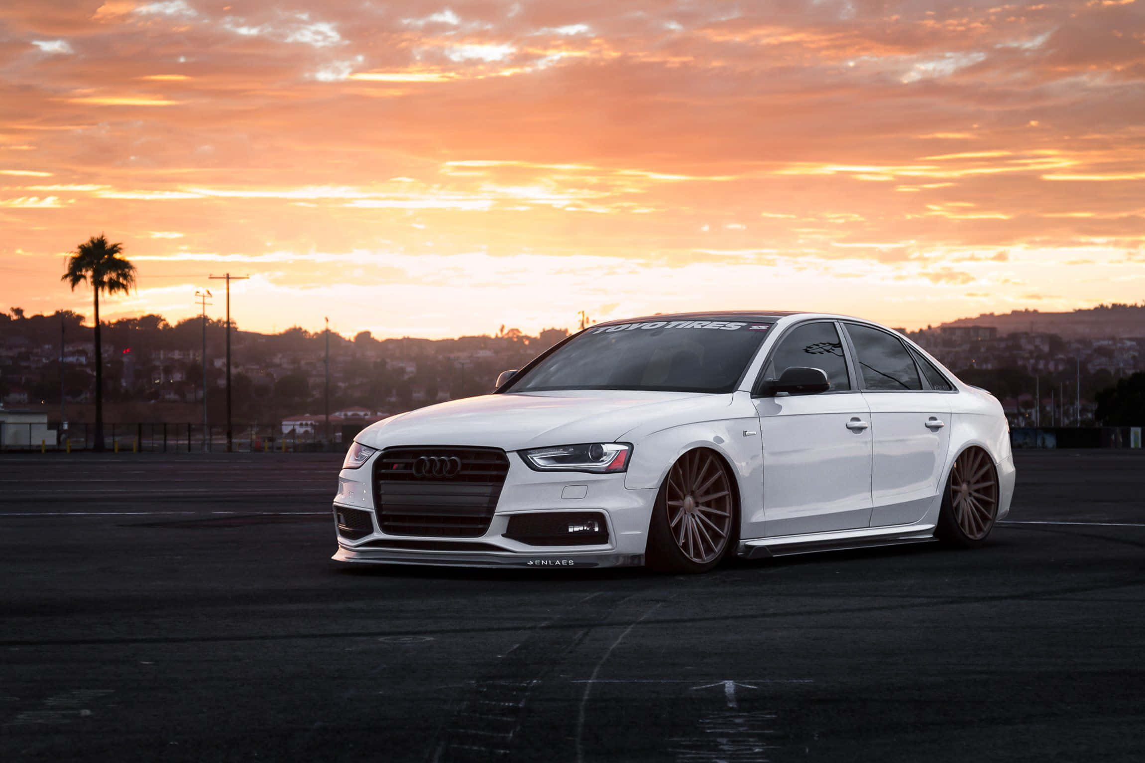 Audi S4 桌布 壁紙 — 一輛時尚的白色奧迪轎車，配備定制青銅輪圈，停在瀝青路面上，背景是火紅的橙色日落天空。