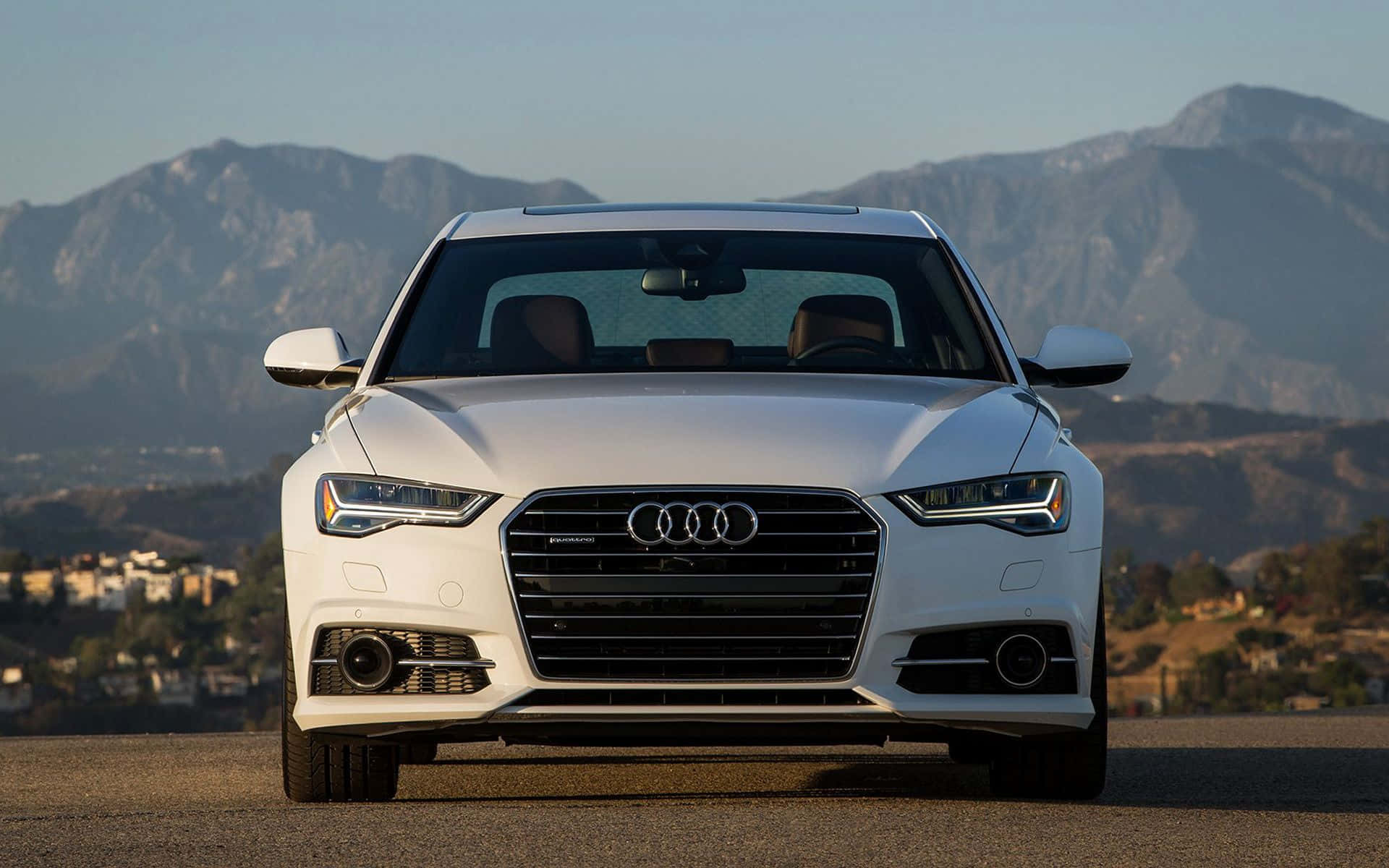 Audi A6 桌布 壁紙 — 一輛時尚的白色奧迪轎車在鋪砌的道路上向前行駛，背景是崎嶇的山脈和晴朗的天空。