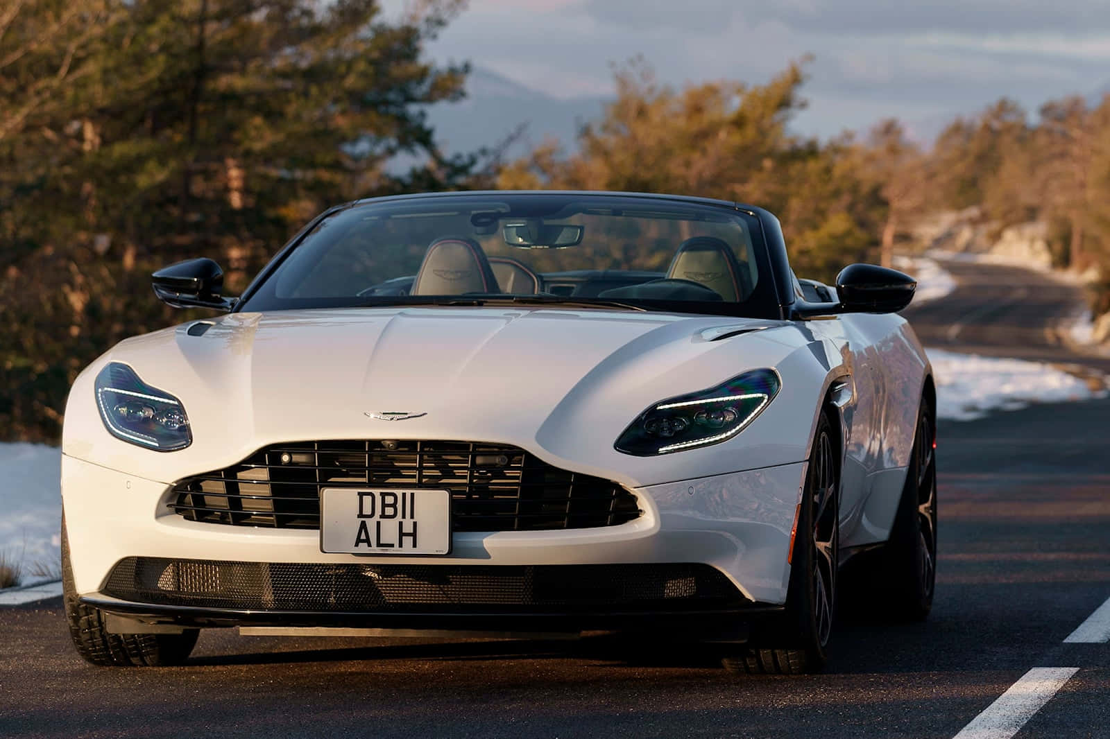 Aston Martin Db11 桌布 壁紙 — 一輛時尚的白色Aston Martin DB11敞篷車停泊在蜿蜒的山路上，周圍有被雪覆蓋的樹木和遠峰，背景是柔和的金色日落。