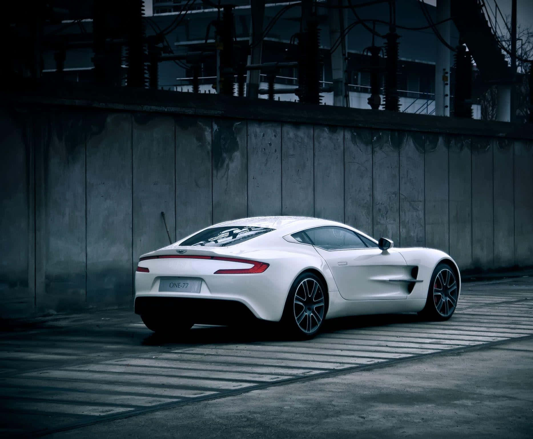 Aston Martin One-77 桌布 壁紙 — 一輛時尚的白色Aston Martin One-77跑車停在混凝土表面上，旁邊是風化的工業牆，光線昏暗。