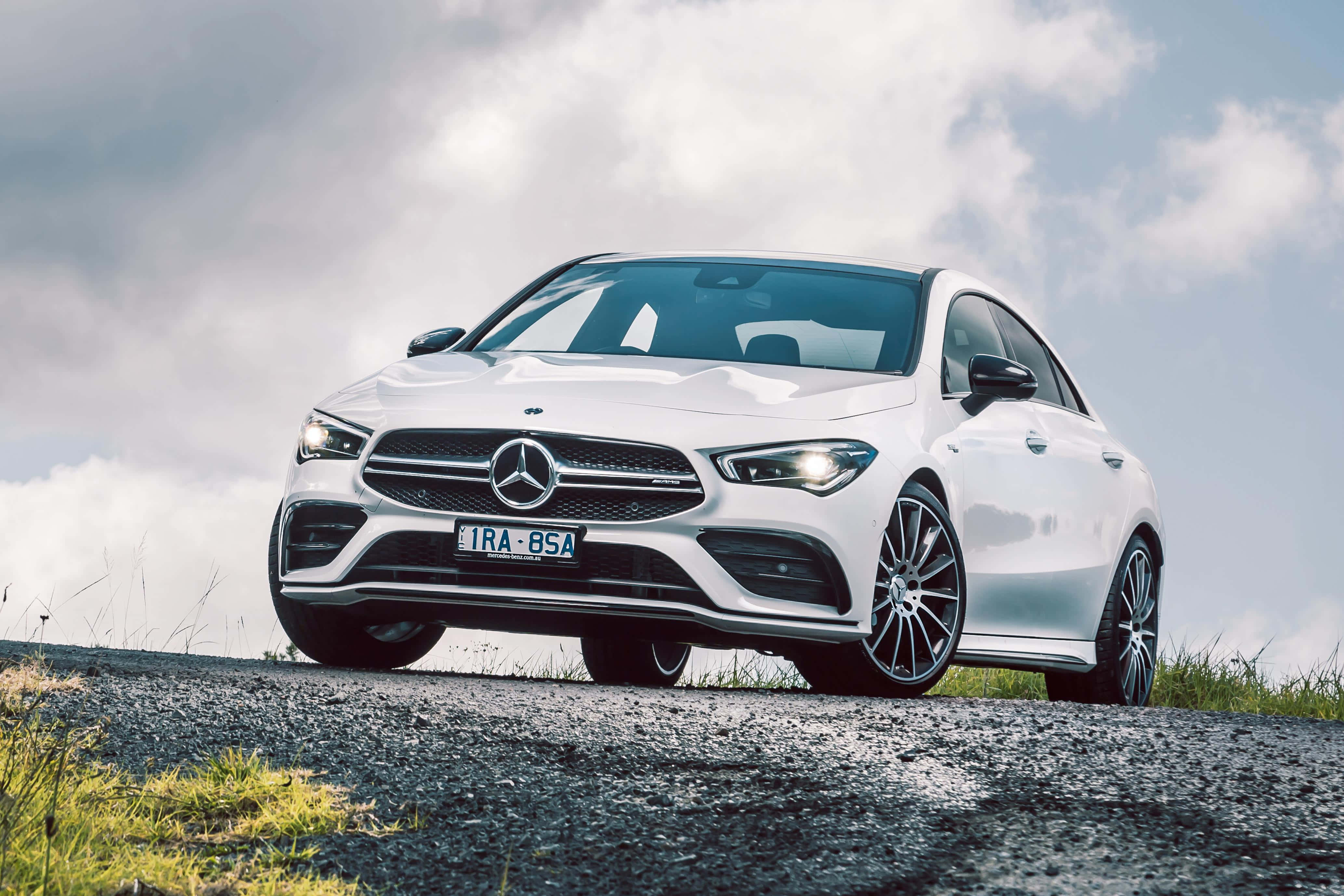 Mercedes Benz Cla-class 桌布 壁紙 — 一輛時尚的白色賓士CLA轎跑車停在碎石路上，天空多雲，路邊有草叢。