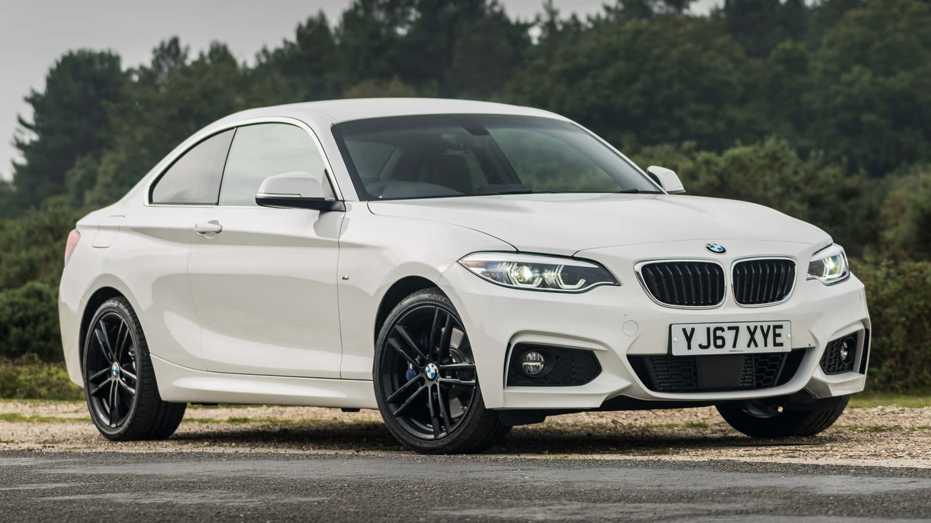 Bmw 2 Series 桌布 壁紙 — 一輛時尚的白色BMW 2系列轎跑車，配黑色輪圈，停在碎石路上，旁邊是樹林，天空多雲。