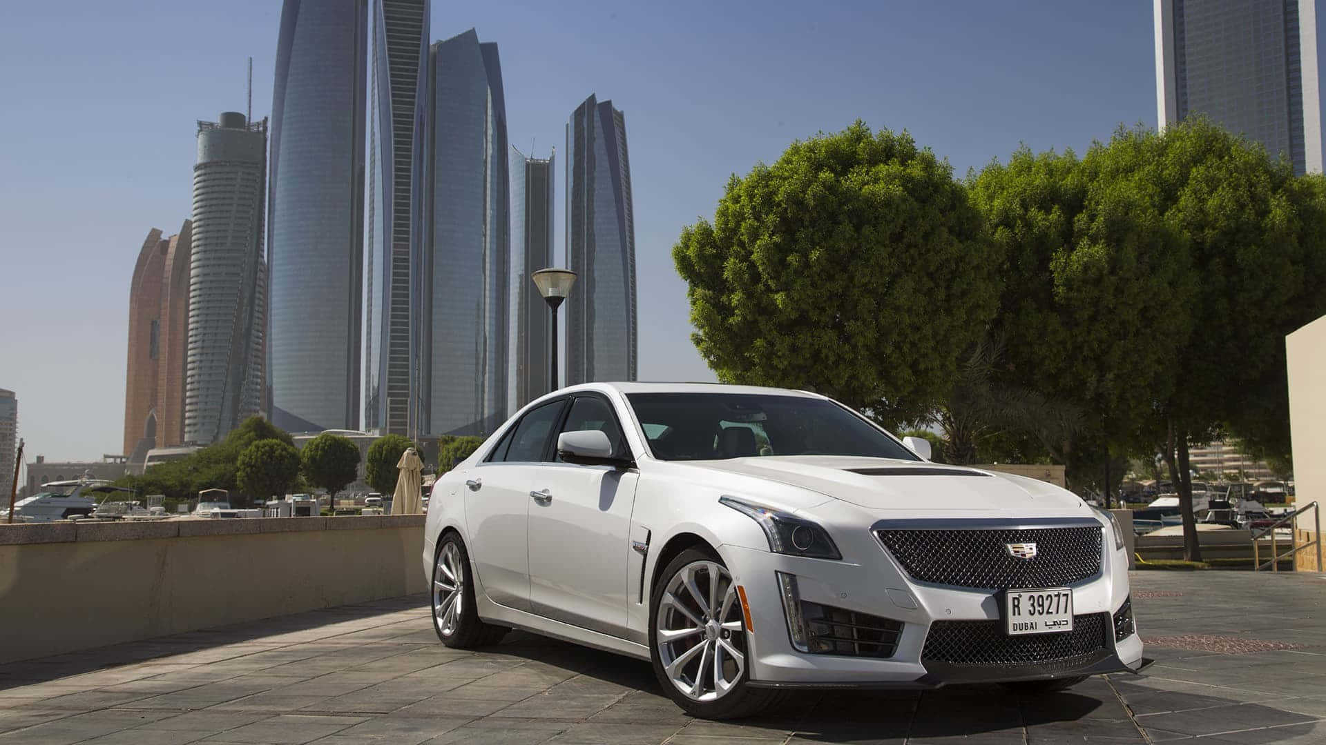 Cadillac Ats 桌布 壁紙 — 一輛時尚的白色Cadillac CTS-V停放在陽光明媚的城市廣場上，背景是未來主義的摩天大樓和茂密的綠色樹木。