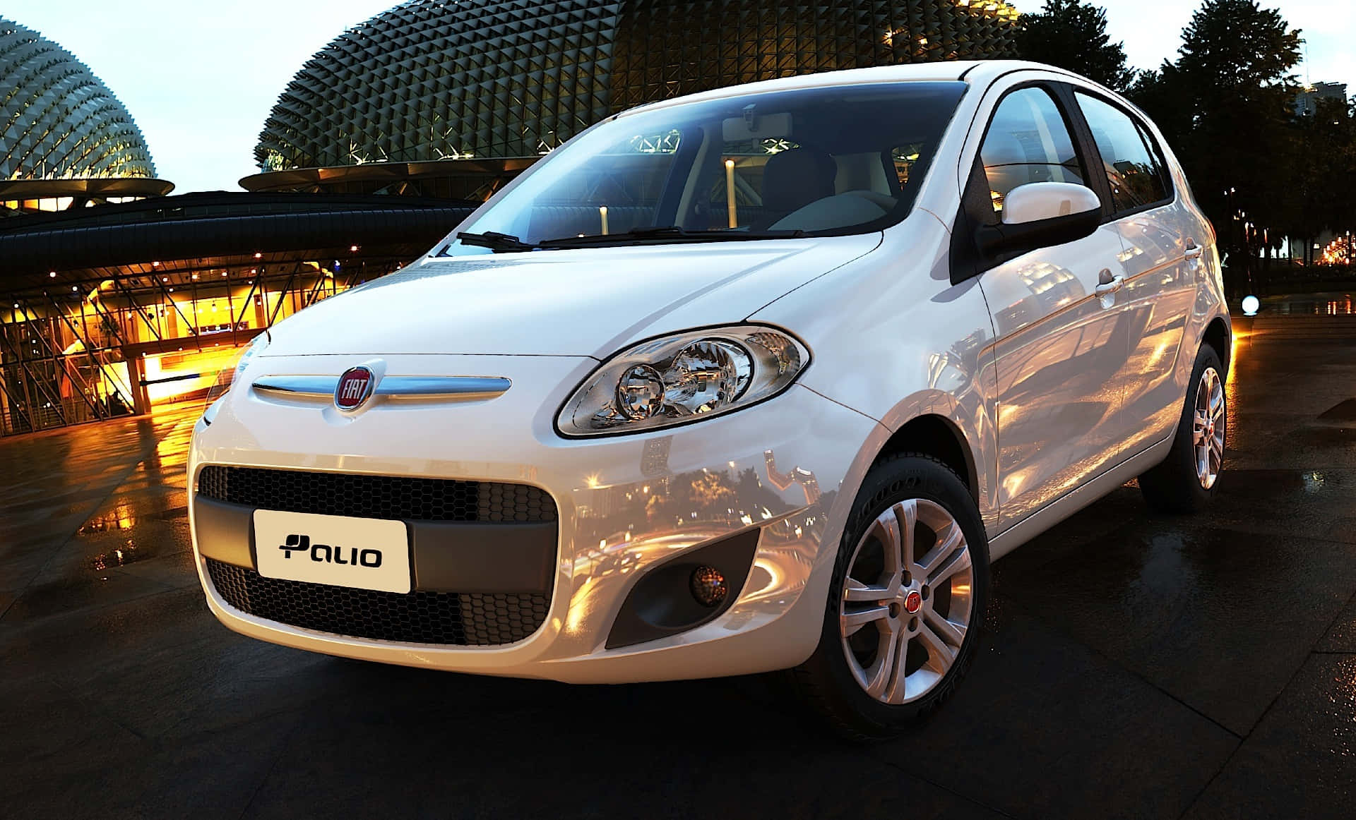 Fiat Palio 桌布 壁紙 — 一輛時尚的白色菲亞特派力奧停泊在黃昏時濕漉漉的路面上，背景是燈火通明的現代圓頂建築。