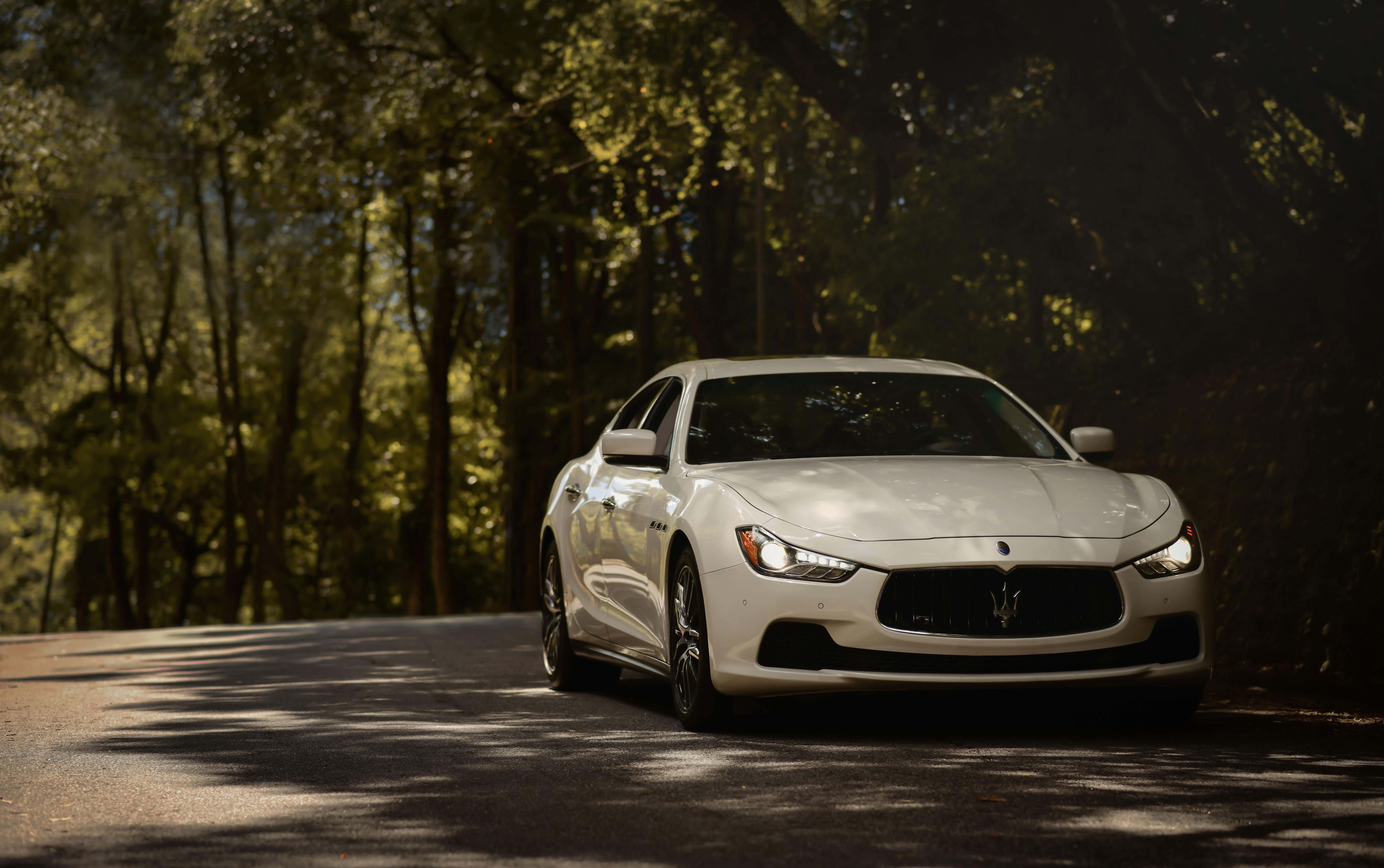 Maserati Ghibli 桌布 壁紙 — 一輛時尚的白色瑪莎拉蒂跑車沿著陽光斑駁的森林道路行駛，高大的樹木投下斑駁的陰影。