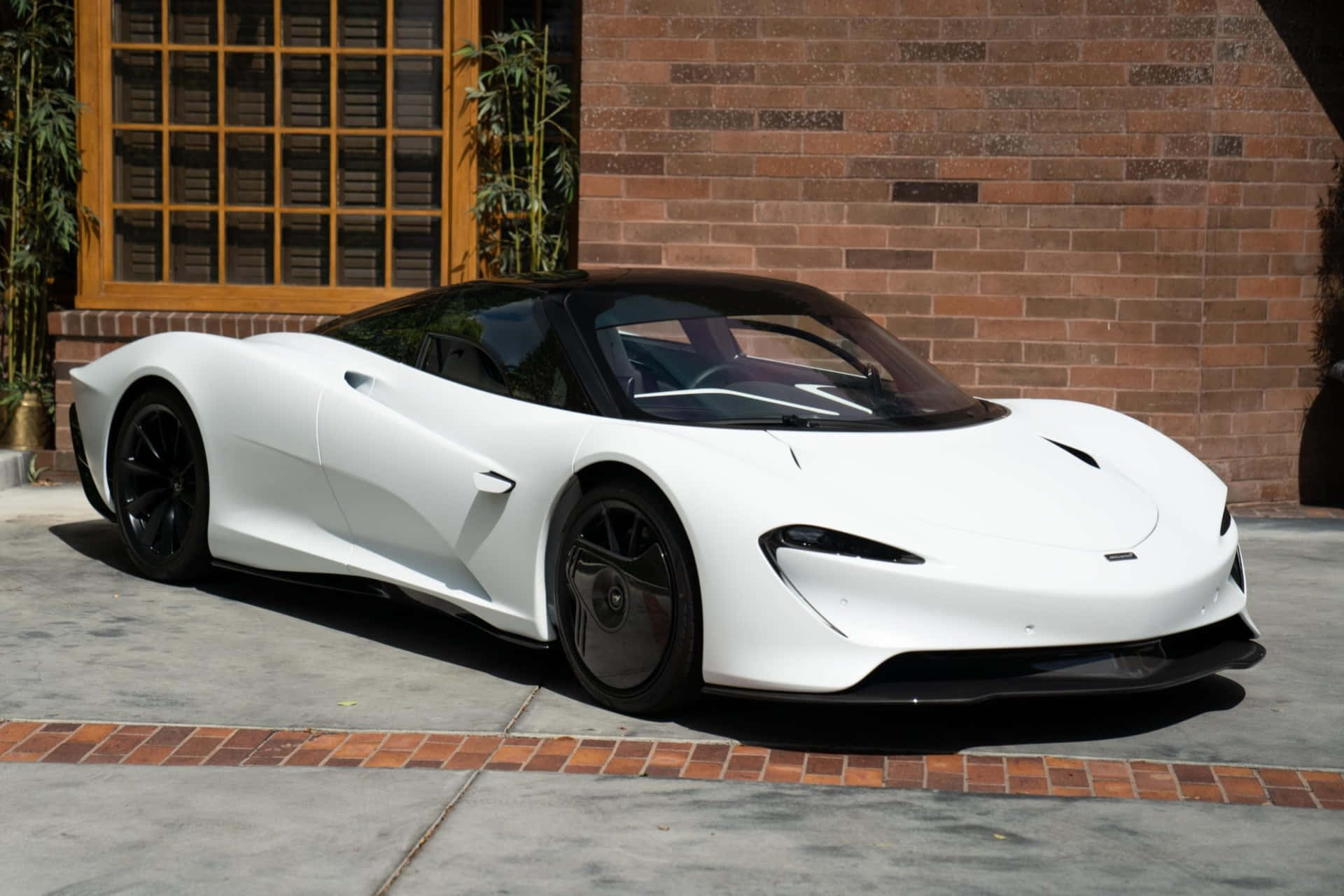 Mclaren Speedtail 桌布 壁紙 — 一輛時尚的白色McLaren Speedtail跑車停在磚牆和竹植物旁邊的混凝土車道上，在明亮的陽光下。
