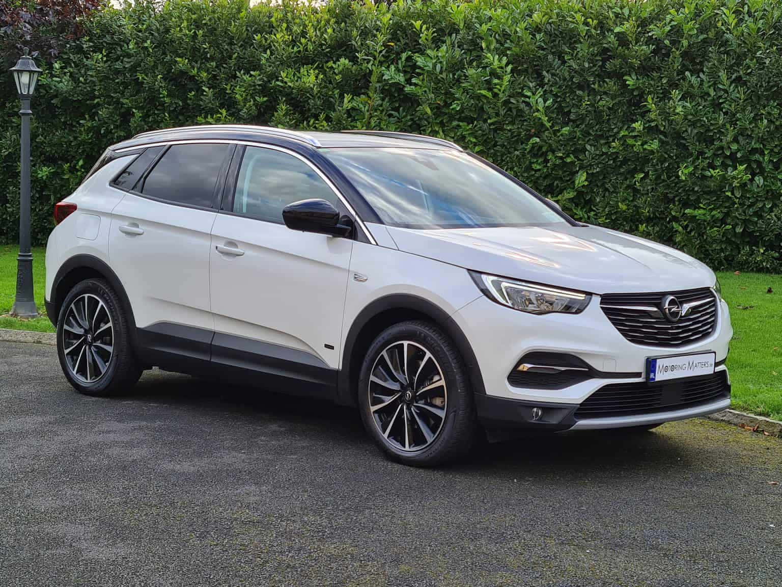 Opel Grandland X 桌布 壁紙 — 一輛時尚的白色Opel SUV停泊在鋪砌的車道上，旁邊是茂密的綠色籬笆和街燈柱。
