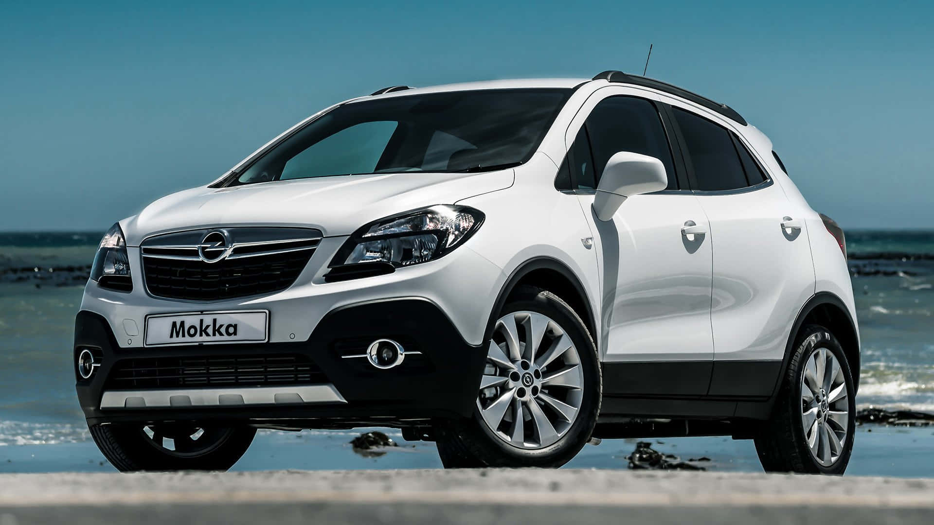 Opel Mokka 桌布 壁紙 — 一輛時尚的白色歐寶 Mokka SUV 停泊在海岸混凝土板上，背後有海浪拍打，天空湛藍清澈。