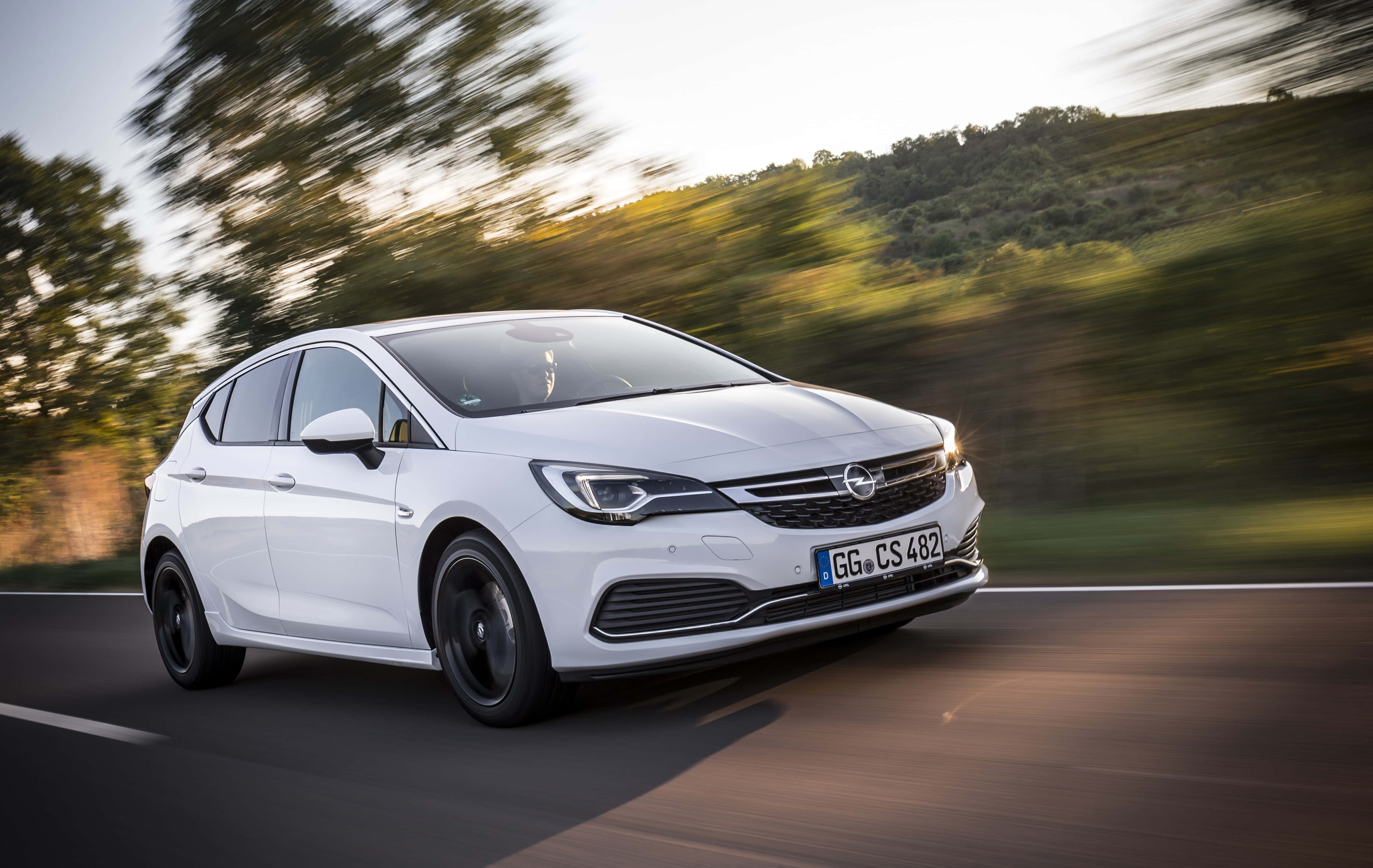 Opel Astra 桌布 壁紙 — 一輛時尚的白色歐寶雅特CS在蜿蜒的道路上行駛，背景是金色光線下模糊的綠色山丘和樹木。