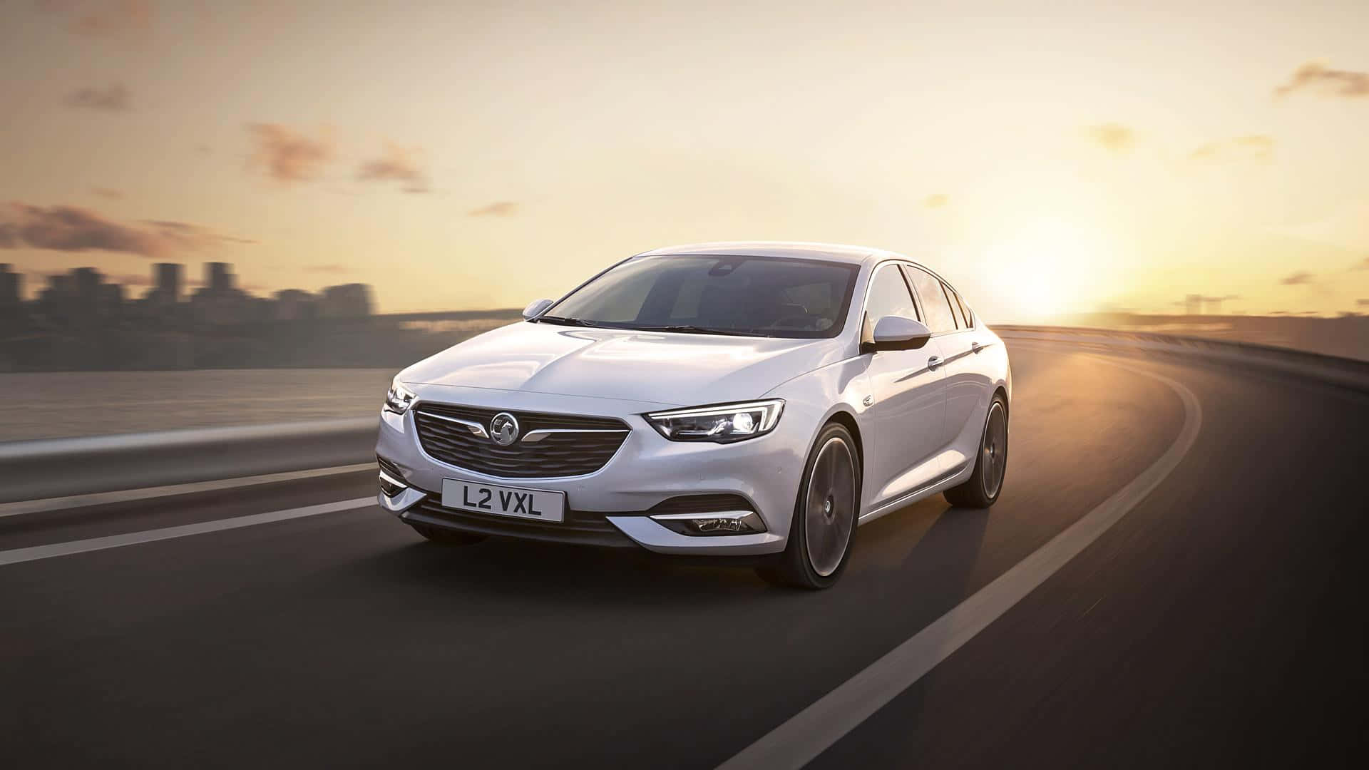 Opel Insignia 桌布 壁紙 — 一輛時尚的白色Vauxhall轎車在日落時分沿著彎曲的高速公路奔馳，遠處是模糊的城市天際線。
