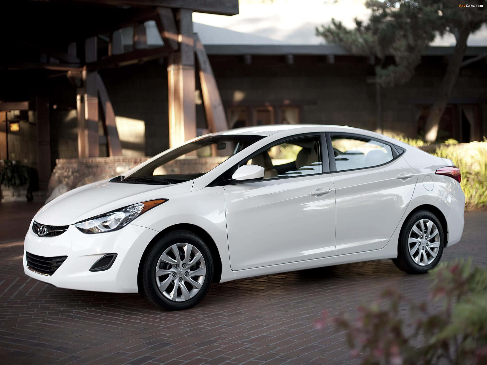 Hyundai Elantra 桌布 壁紙 — 一輛時尚的白色現代伊蘭特轎車停放在磚砌車道上，前方是一座現代木造建築，柔和陽光投射出溫暖陰影。
