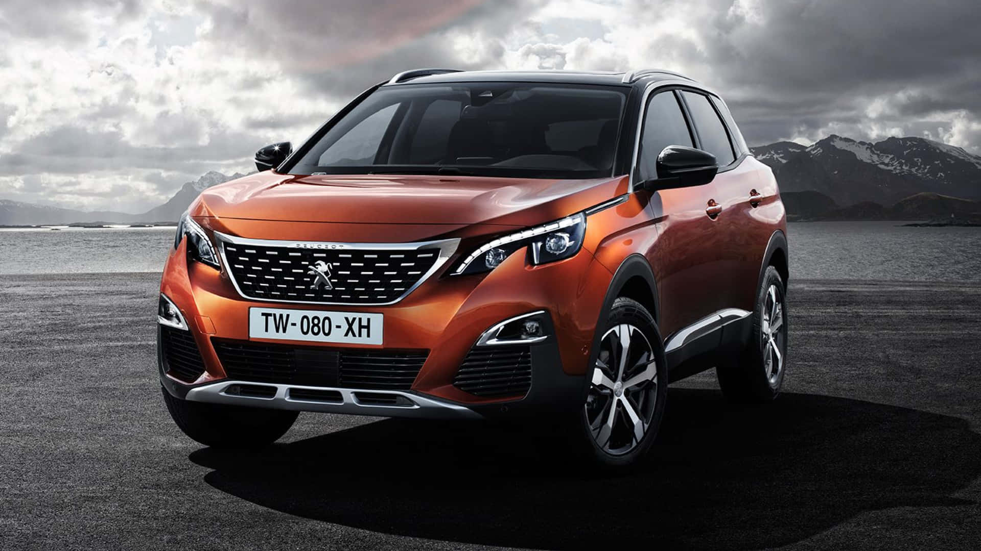  壁紙 — 一輛時尚的橙色Peugeot SUV停在深色瀝青路面上，背景是白雪覆蓋的山脈和多雲的天空。