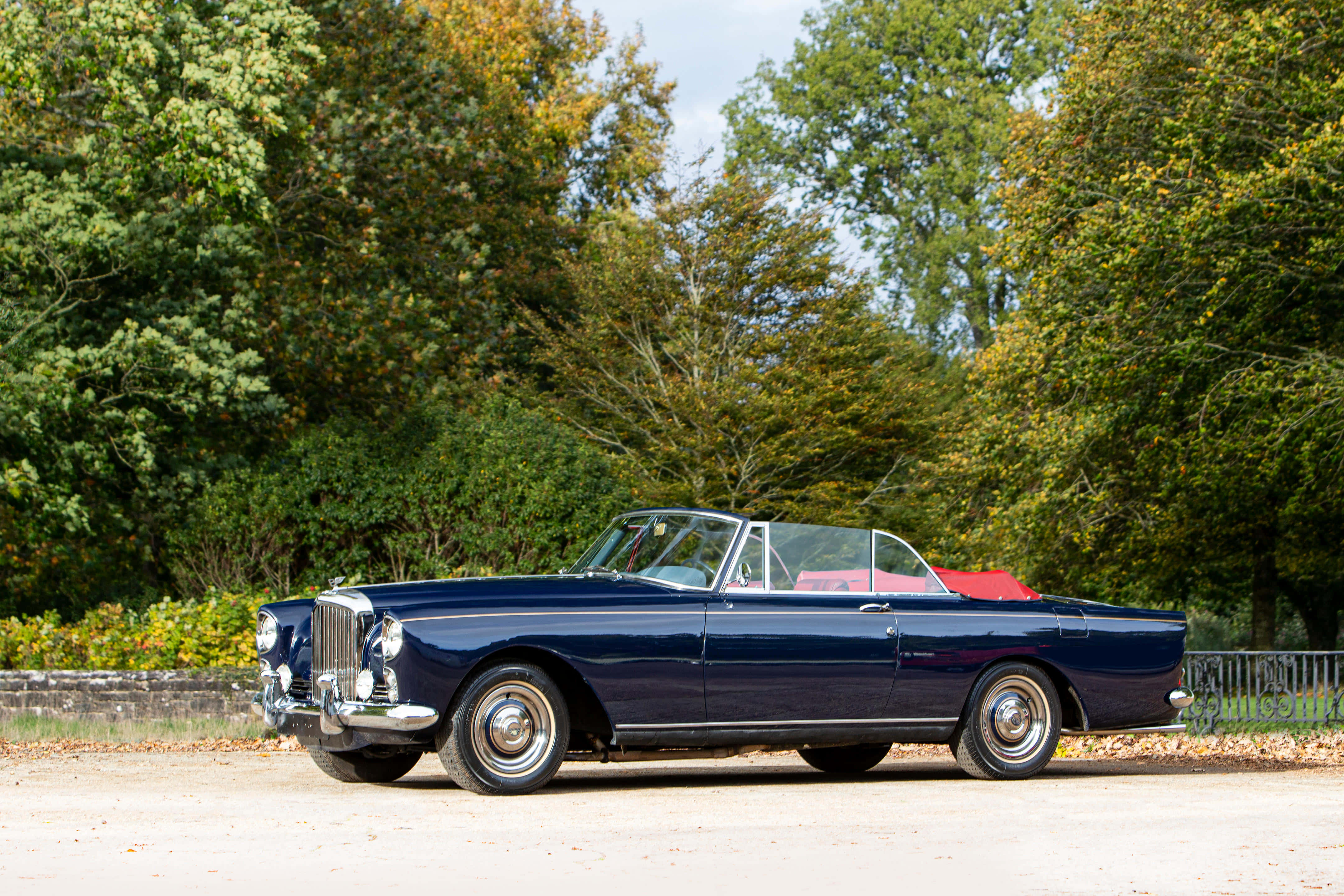 Bentley S2 桌布 壁紙 — 一輛時尚的海軍藍經典敞篷車，紅色軟頂，停在鋪滿碎石的小路上，周圍是秋天的樹木。