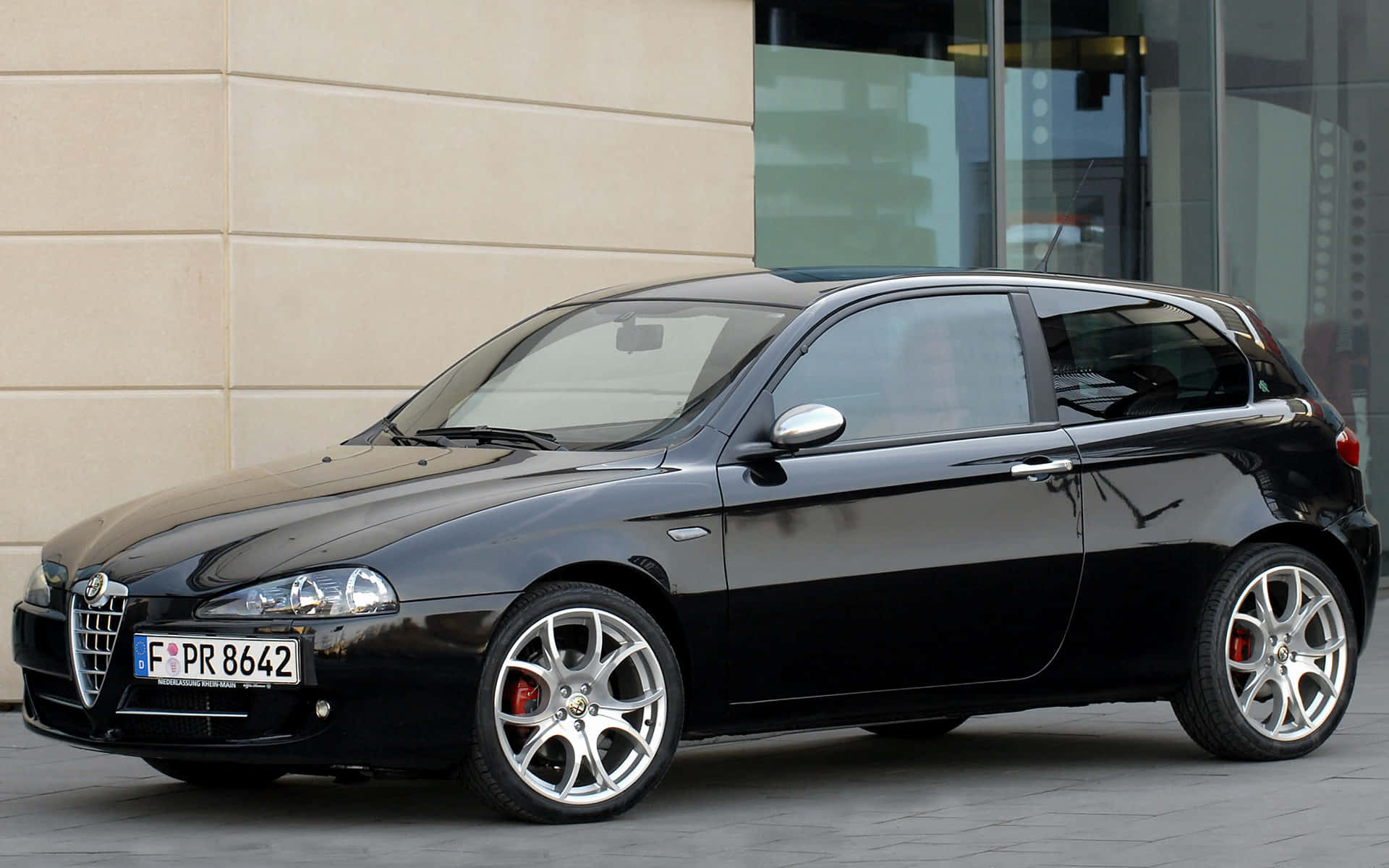 Alfa Romeo 147 桌布 壁紙 — 一輛時尚的黑色愛快羅密歐147跑車停泊在鋪砌的城市街道上，旁邊是一座帶玻璃窗的現代建築。
