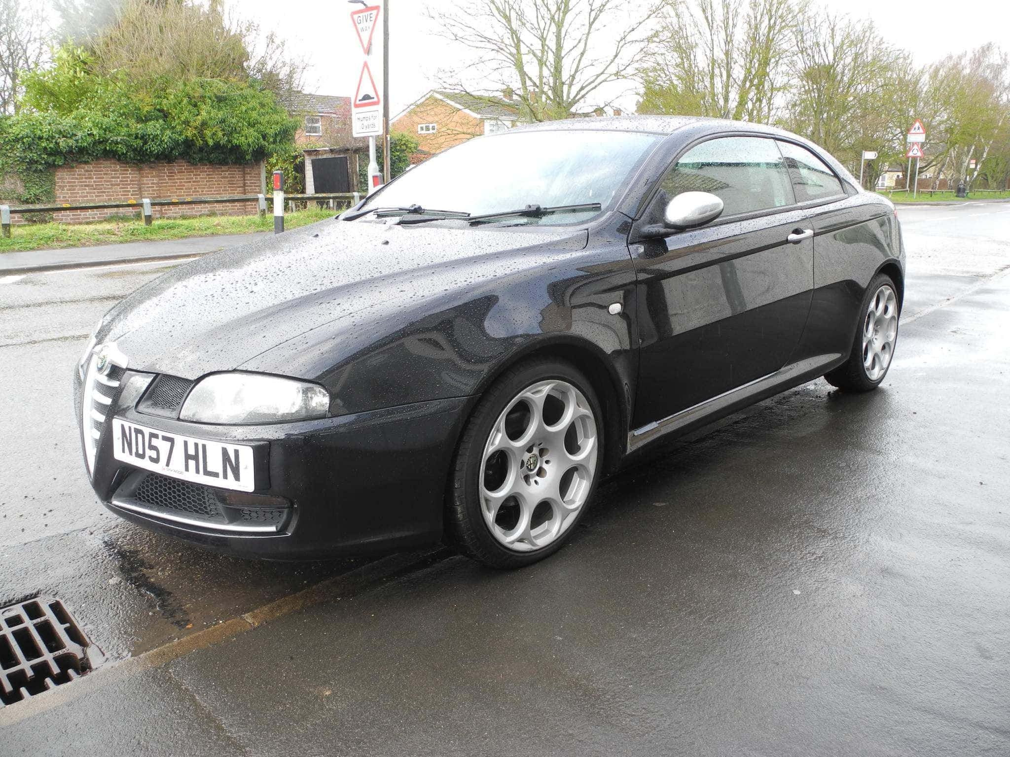 Alfa Romeo Gt 桌布 壁紙 — 一輛時尚的黑色愛快羅密歐轎跑車停在潮濕的住宅街道上，天空多雲。
