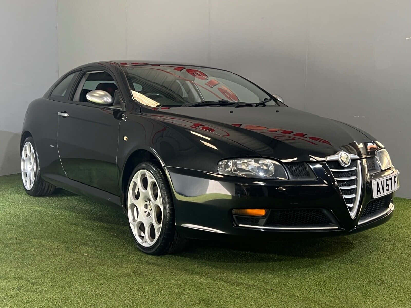 Alfa Romeo Gt 桌布 壁紙 — 一輛時尚的黑色愛快羅密歐雙門轎跑車，配備銀色輪圈，停放在綠色草地上，背景是純灰色牆壁。