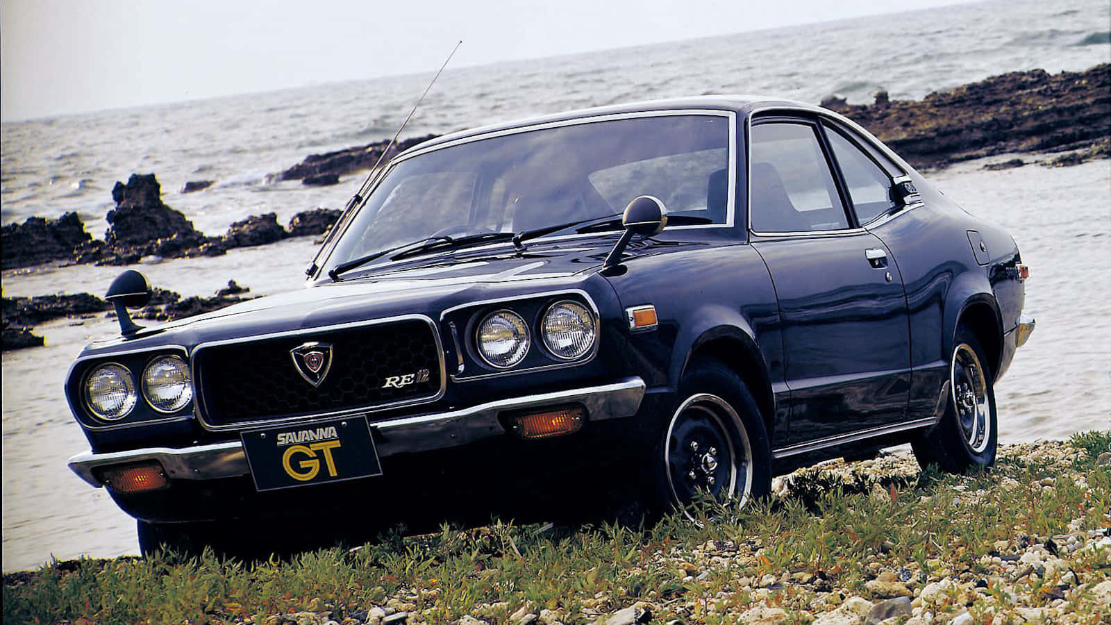 Mazda Rx-3 桌布 壁紙 — 一輛時尚的黑色Datsun 510 GT轎跑車停泊在岩石海邊懸崖上，俯瞰著洶湧的海浪。