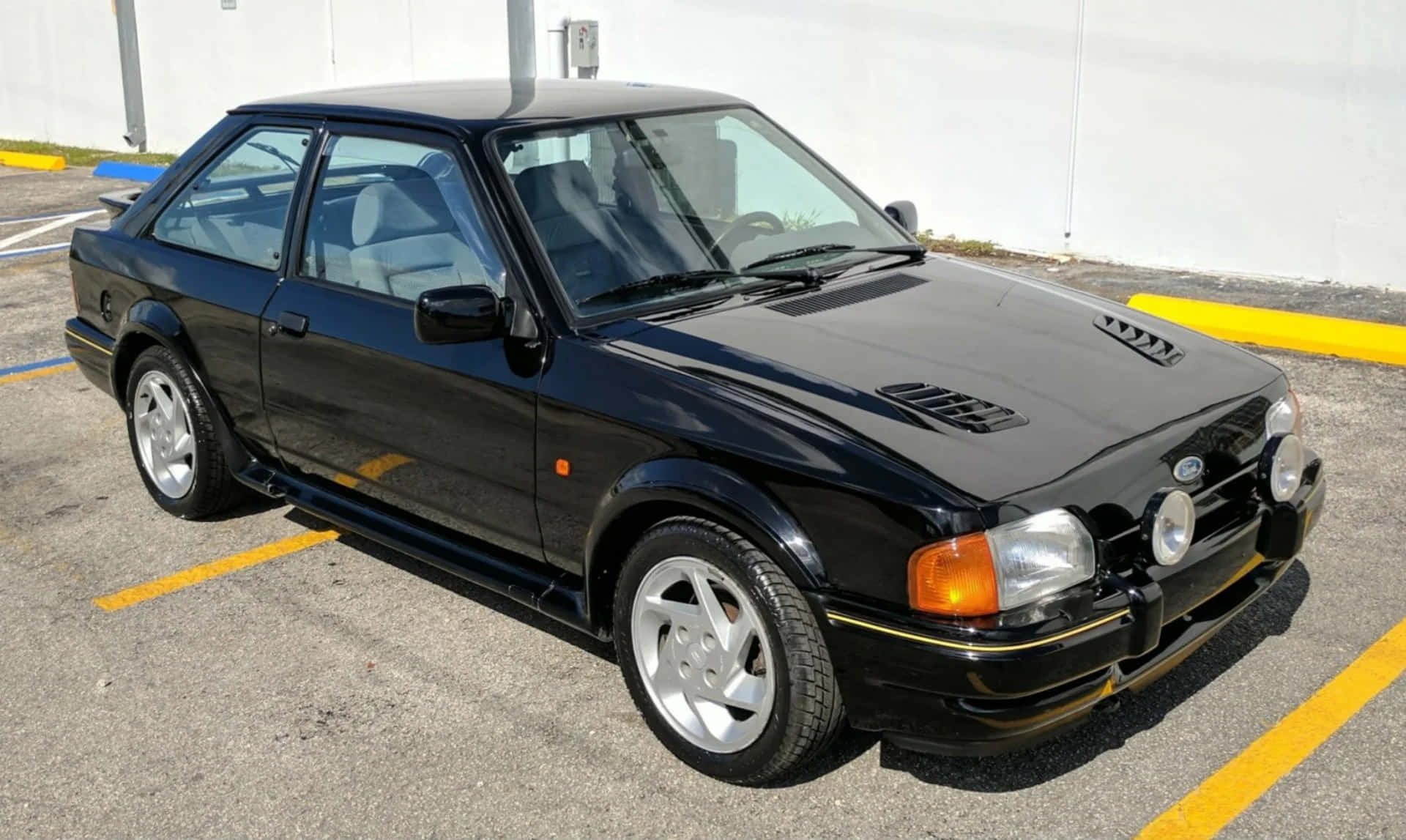Ford Escort 桌布 壁紙 — 一輛時尚的黑色福特Sierra Cosworth跑車停放在陽光普照的瀝青停車場，地面有黃色標線。
