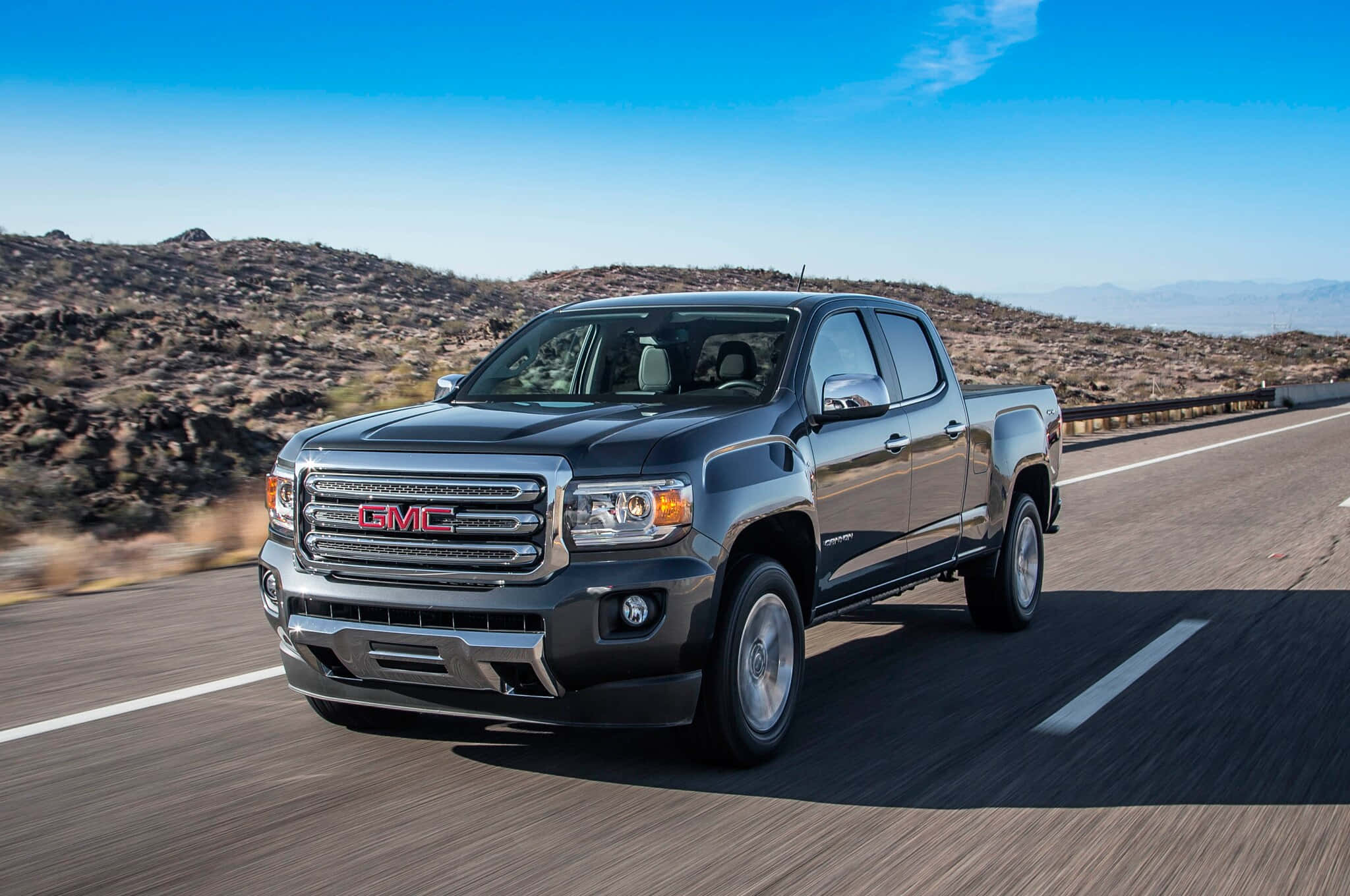 Gmc Canyon 桌布 壁紙 — 一輛時尚的黑色GMC Canyon皮卡車在晴朗藍天下，沿著沙漠高速公路疾馳，背景是岩石丘陵。