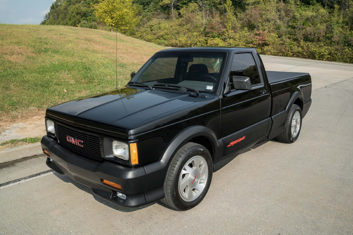 Gmc Syclone 桌布 壁紙 — 一輛時尚的黑色GMC Syclone pickup貨車停在鋪設的道路上，旁邊是長滿秋樹的草坡。