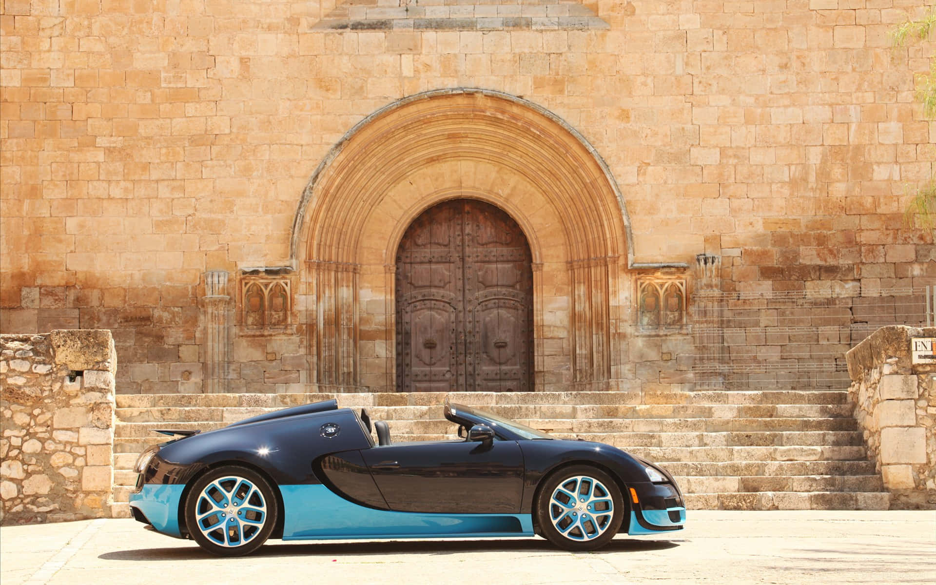Bugatti Veyron 桌布 壁紙 — 一輛時尚的黑色和藍色Bugatti Veyron敞篷車停在歷史悠久的石頭教堂前，教堂有拱形木門。