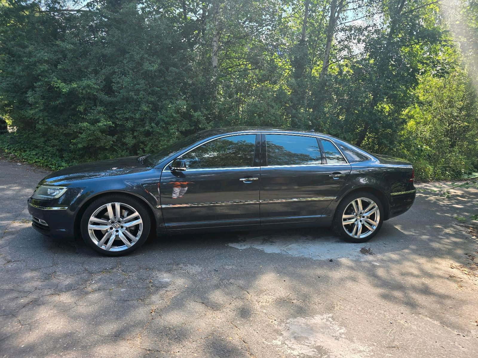 Volkswagen Phaeton 桌布 壁紙 — 一輛時尚的黑色轎車停泊在陽光斑駁的碎石車道上，周圍環繞著茂密的綠樹。