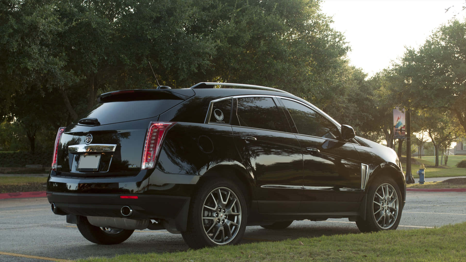 Cadillac Srx 桌布 壁紙 — 一輛時尚的黑色凱迪拉克SUV停在黃金時段樹木成蔭的街道旁的草地上。
