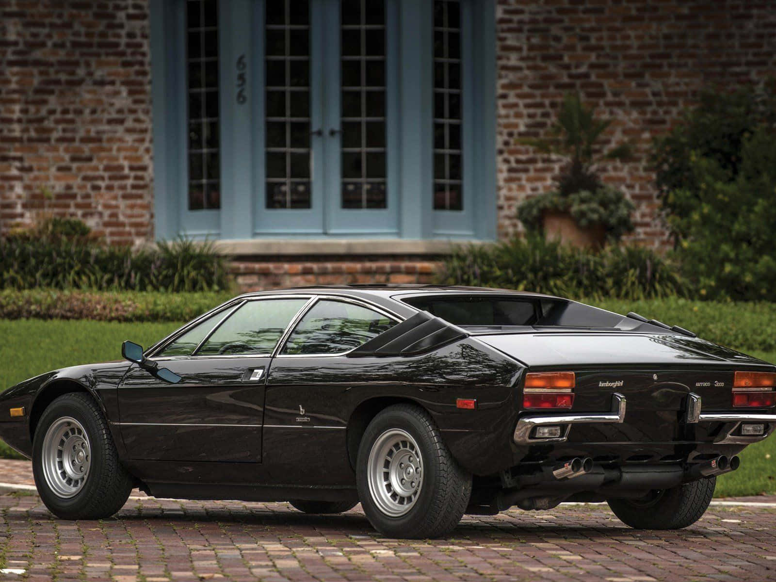 Lamborghini Urraco 桌布 壁紙 — 一輛時尚的黑色藍寶堅尼Countach停在磚砌車道上，前方是間有藍色門的磚屋。