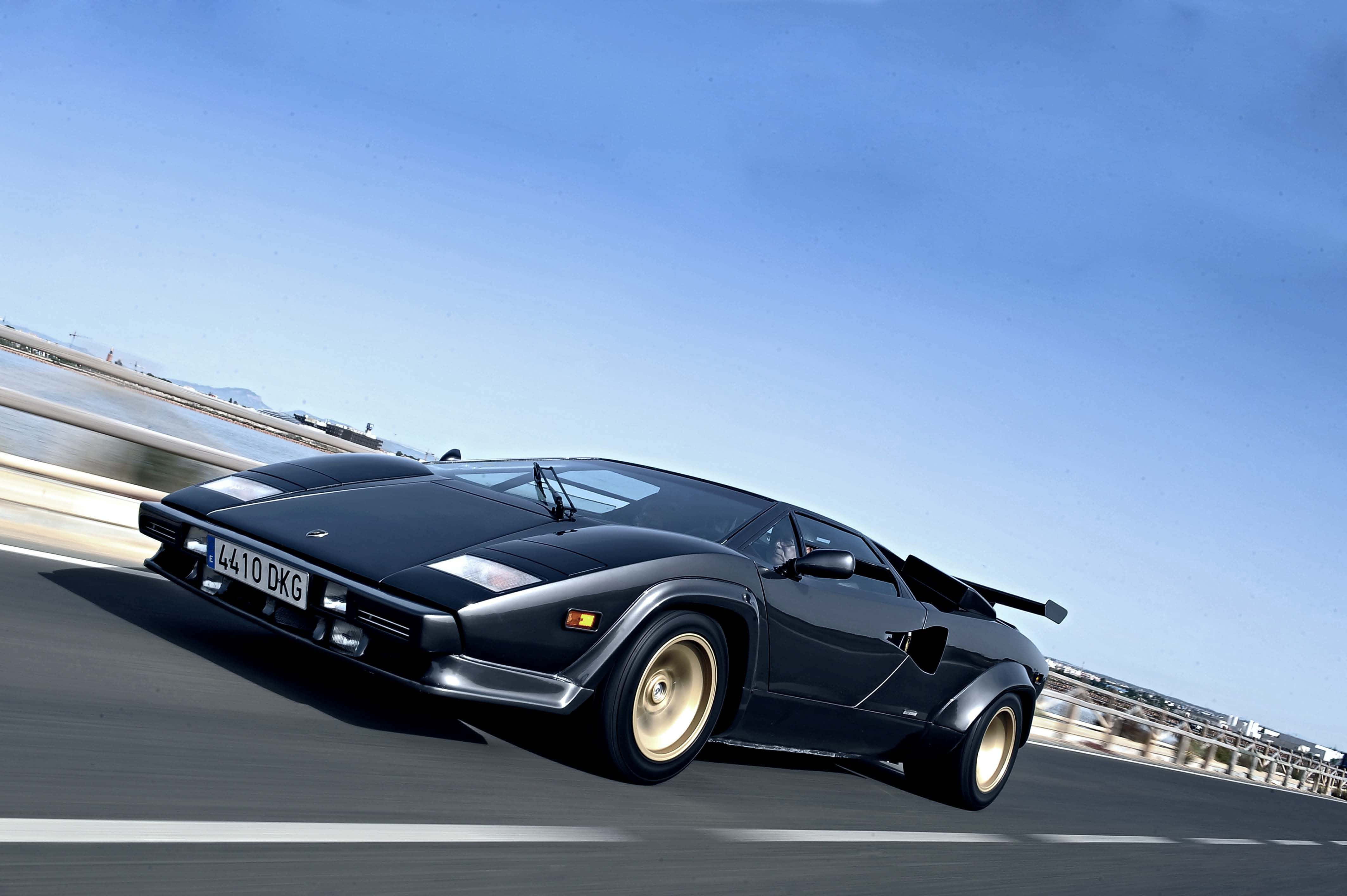 Lamborghini Countach 桌布 壁紙 — 一輛時尚的黑色藍寶堅尼Countach在晴朗的藍天下沿著海岸公路奔馳，動態模糊強調了它的速度。